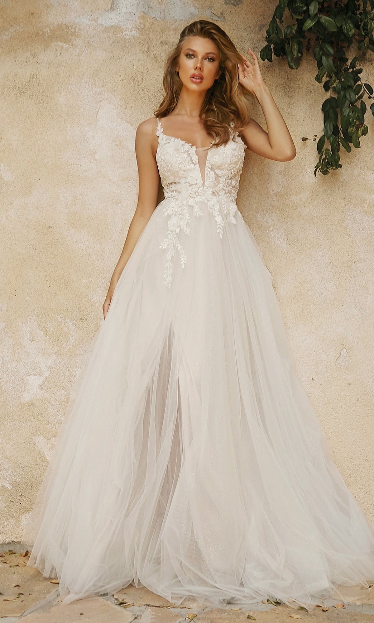 A woman in the Classic Off-White Long Prom Dress (Ladivine CB072W) with floral lace details stands by a beige textured wall, green leaves above her.