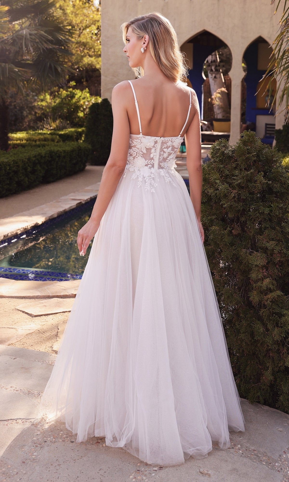 A woman stands outdoors by a pool, wearing the Ladivine CB072W Classic Off-White Long Prom Dress, featuring a sleeveless design, sheer back, and floral lace details.