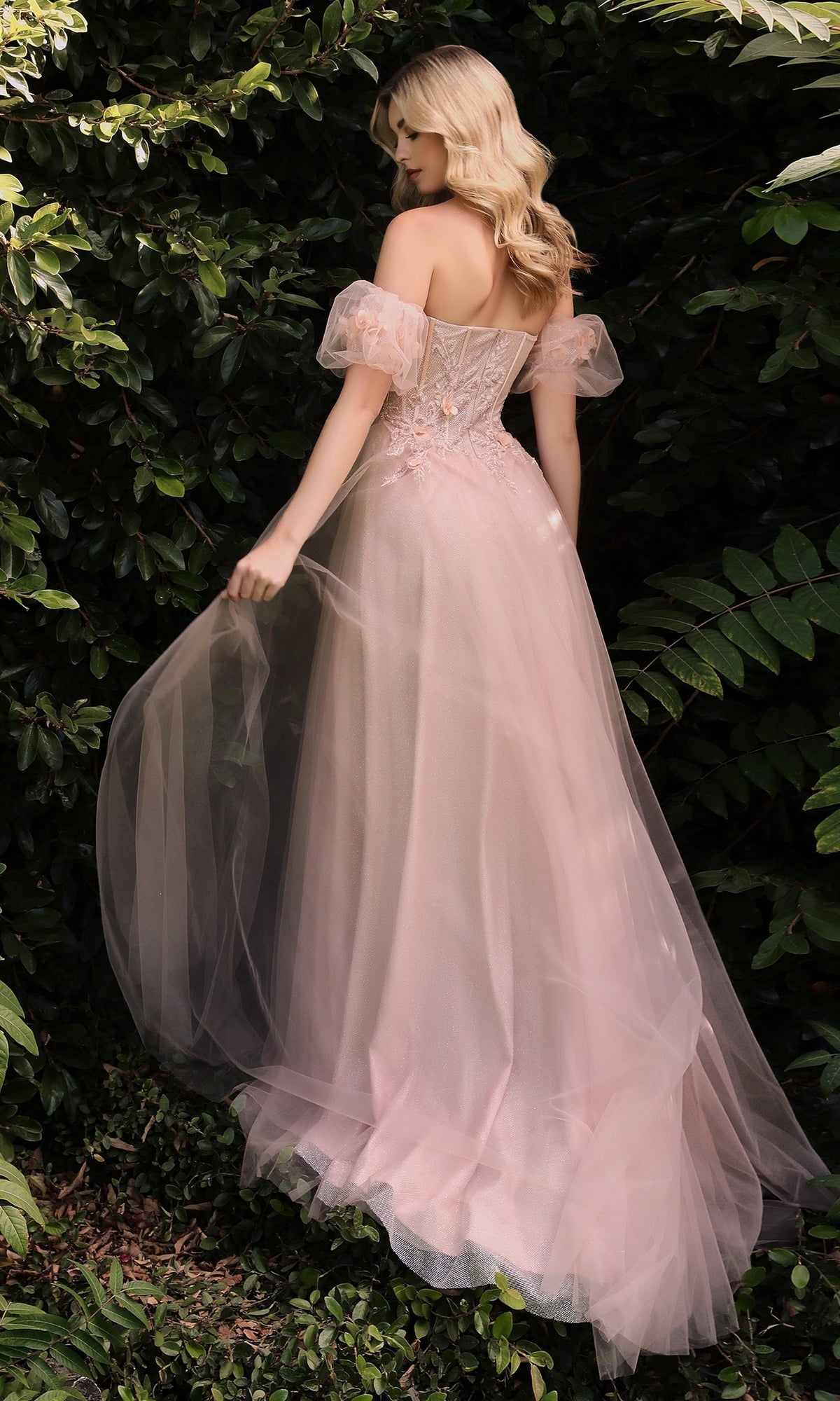A woman in the Ladivine CB080 Glitter Tulle Ball Gown Prom Dress stands outdoors with her back turned, surrounded by lush green foliage.