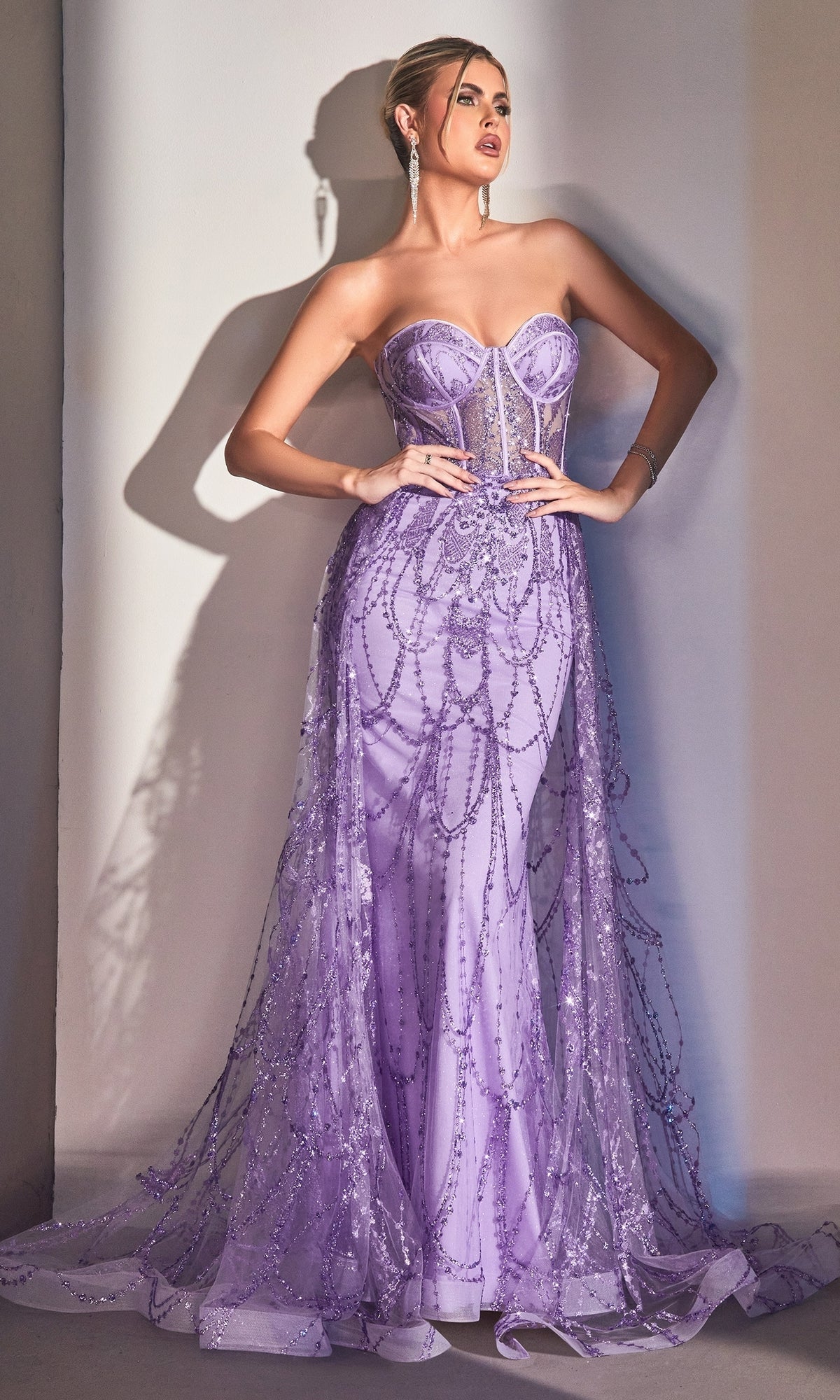 A woman stands elegantly against a white wall wearing the Strapless Mermaid Prom Dress with Overskirt CB095 in lavender, featuring intricate beading and a long, sheer embellished train.