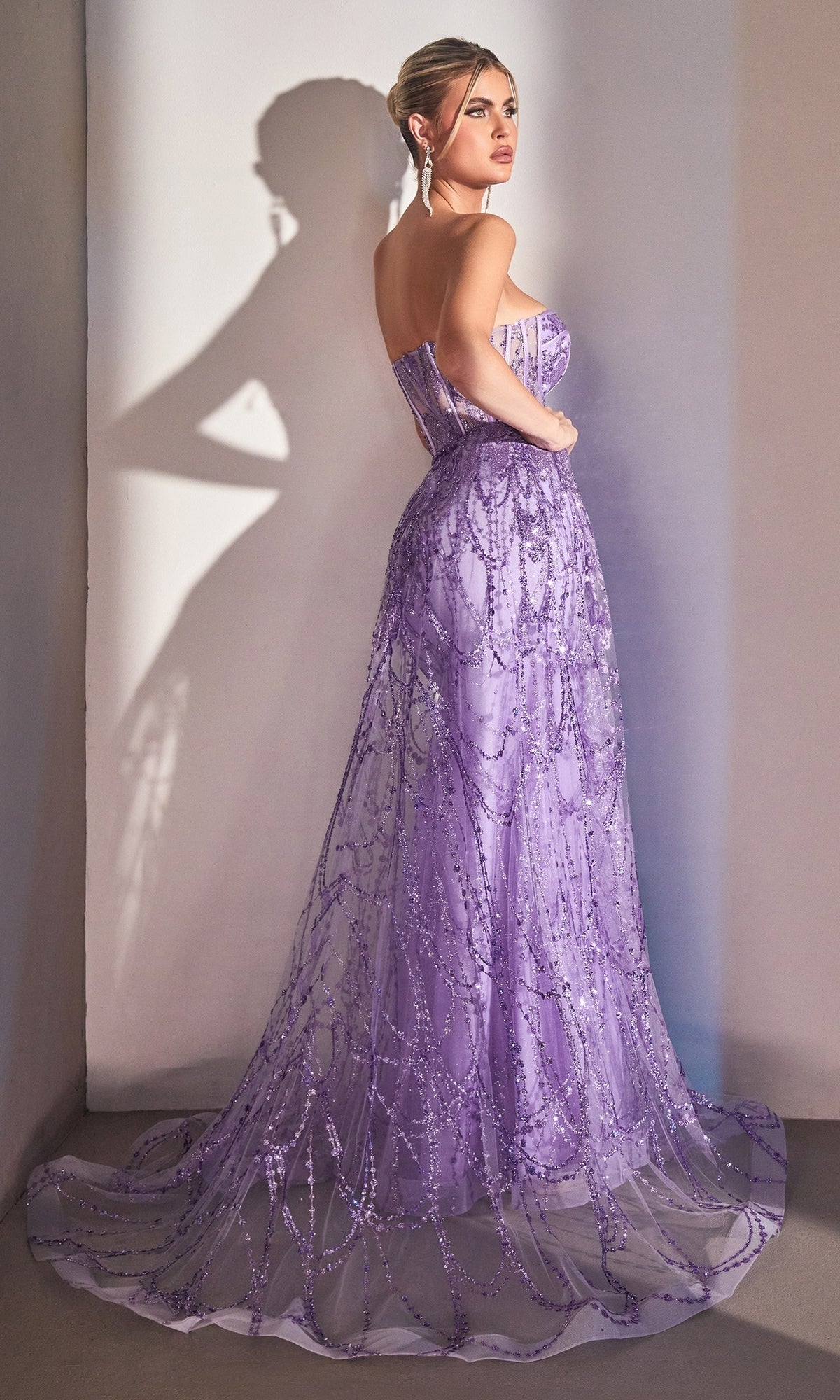 A woman stands against a light wall in the Strapless Mermaid Prom Dress with Overskirt CB095, a floor-length lavender gown with beaded embellishments, creating an elegant and sophisticated evening look.