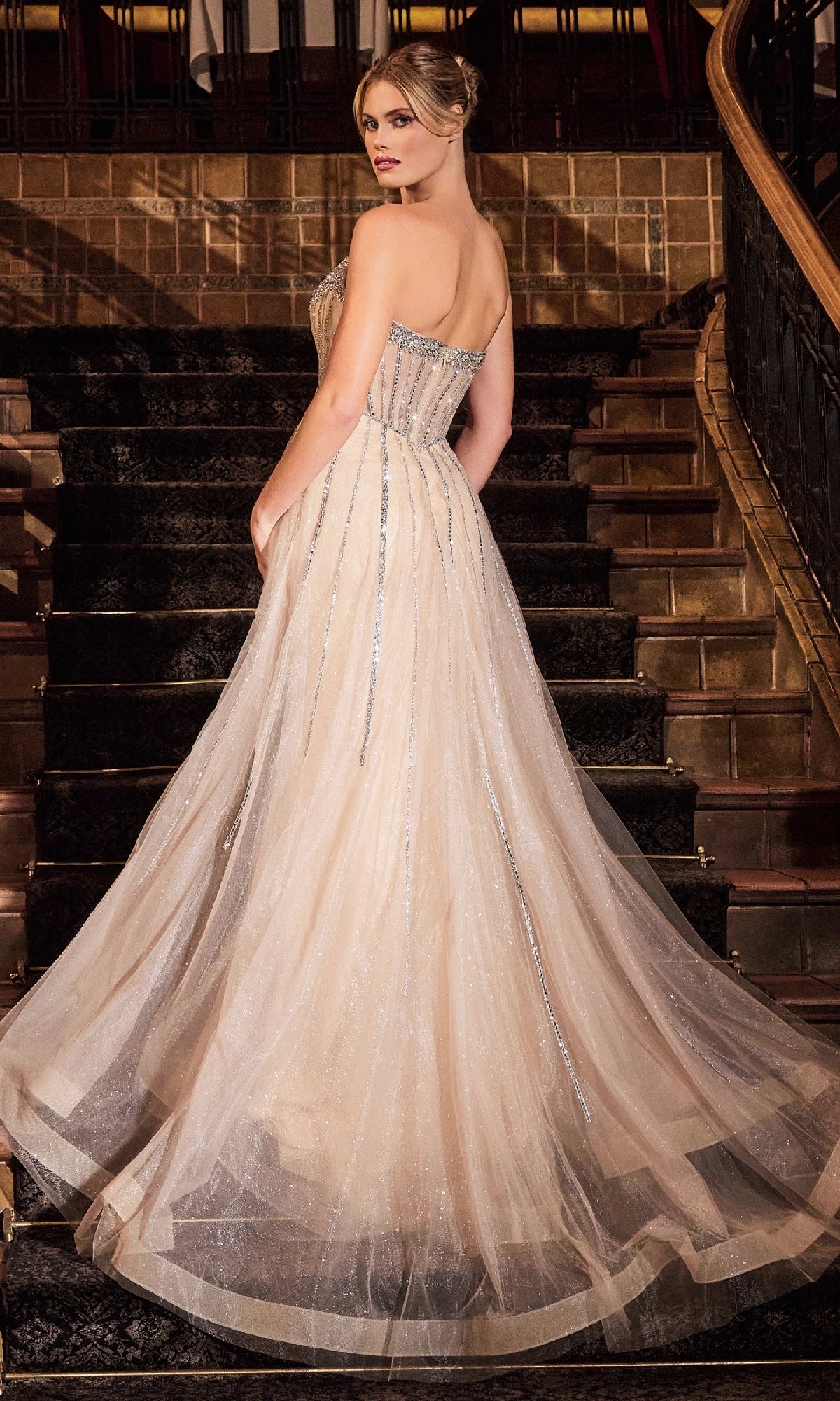A woman in the Strapless Glitter-Tulle Prom Gown: Ladivine CB158, a floor-length cream dress with a fitted, embellished bodice, stands on a staircase and looks over her shoulder—a stunning vision in this elegant formal gown.