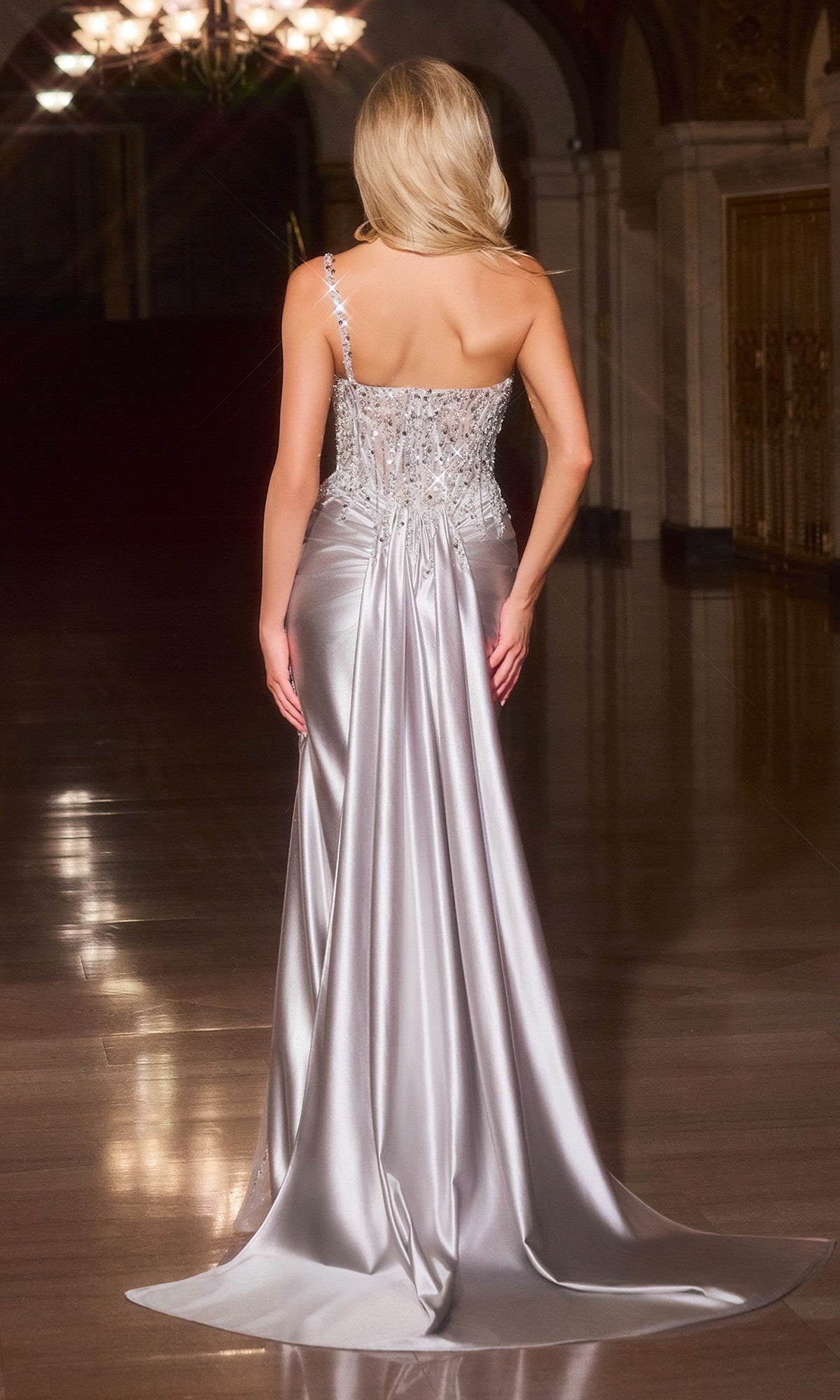 A woman with long blonde hair, wearing the Ladivine CC0329 long prom dress with a sheer corset bodice, stands with her back to the camera in an elegant, dimly lit hall.