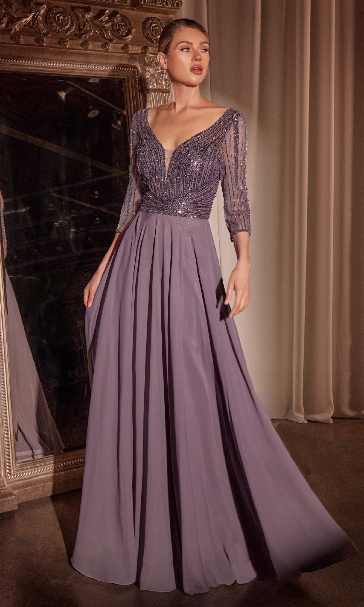 A woman wearing the Modest Long A-Line Formal Dress: Ladivine CC446, featuring a floor-length lavender skirt, beaded fitted bodice, and sheer sleeves, stands near a large ornate mirror.