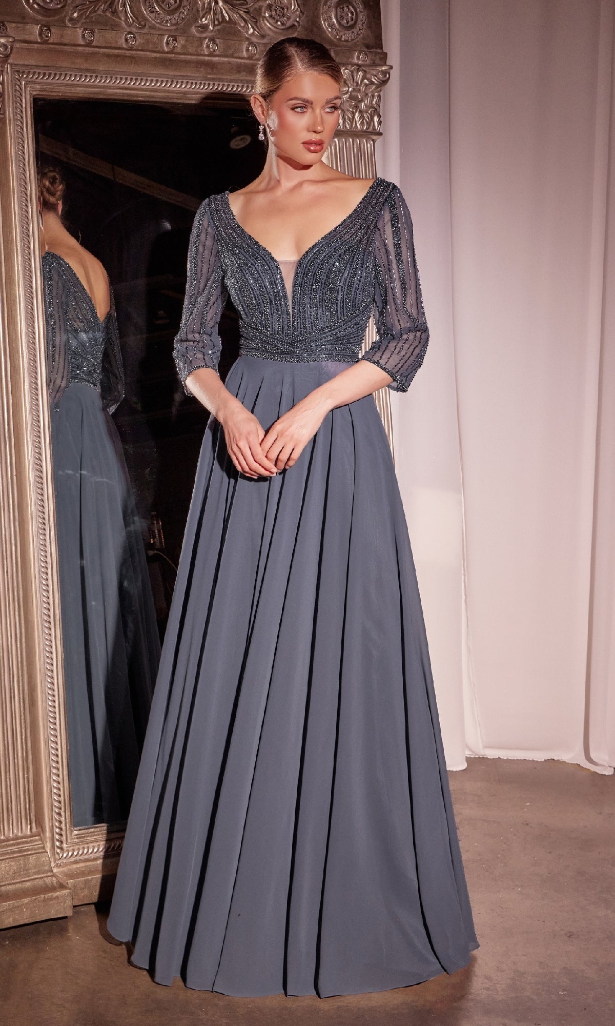 A woman in the Modest Long A-Line Formal Dress: Ladivine CC446, featuring a floor-length slate blue design with sheer three-quarter sleeves and a deep V-neckline, stands before a large ornate mirror.