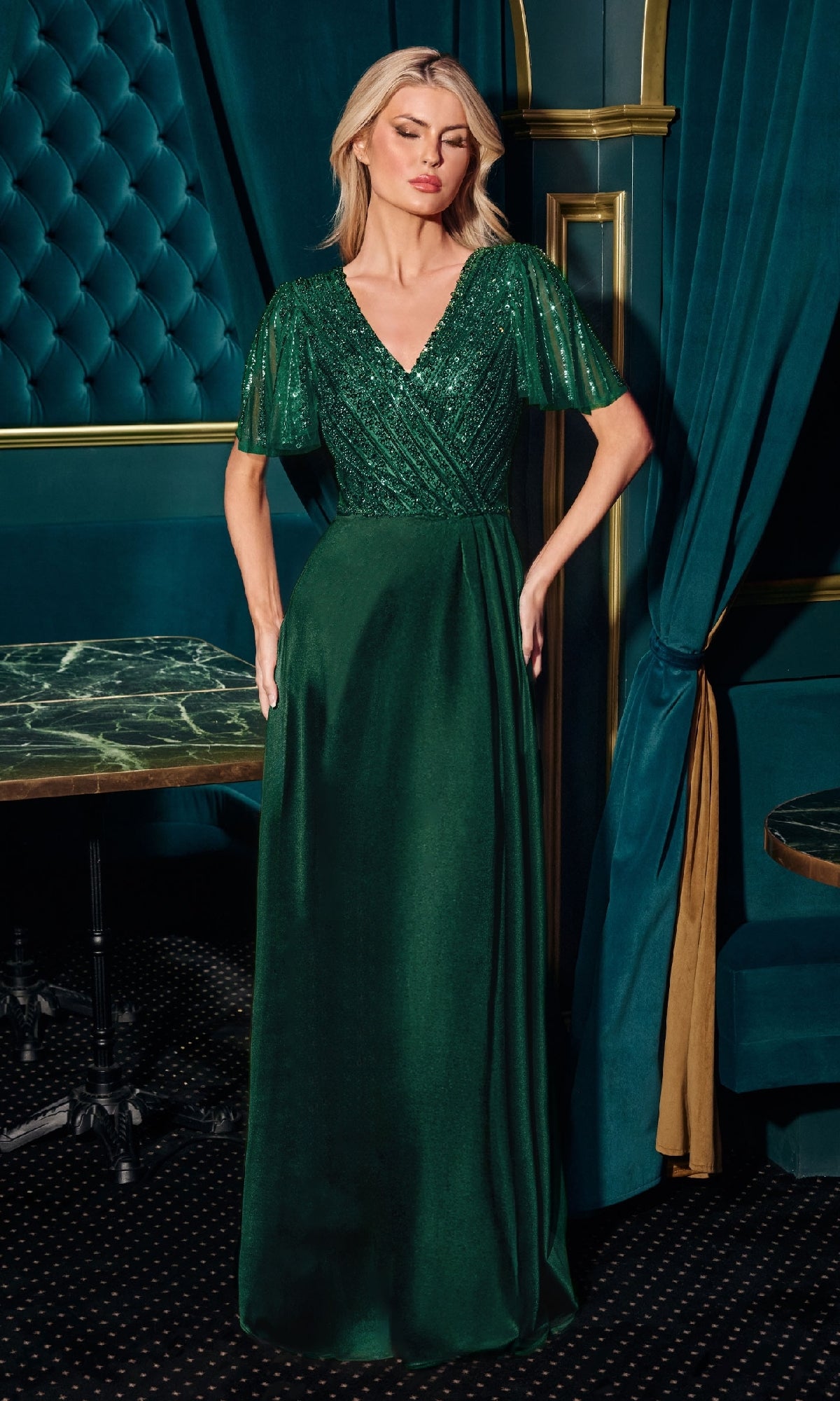 A woman stands indoors wearing the Flutter-Sleeve Long Formal Dress: Ladivine CC447, an emerald green, floor-length gown with sheer flutter sleeves and a fitted embellished bodice.