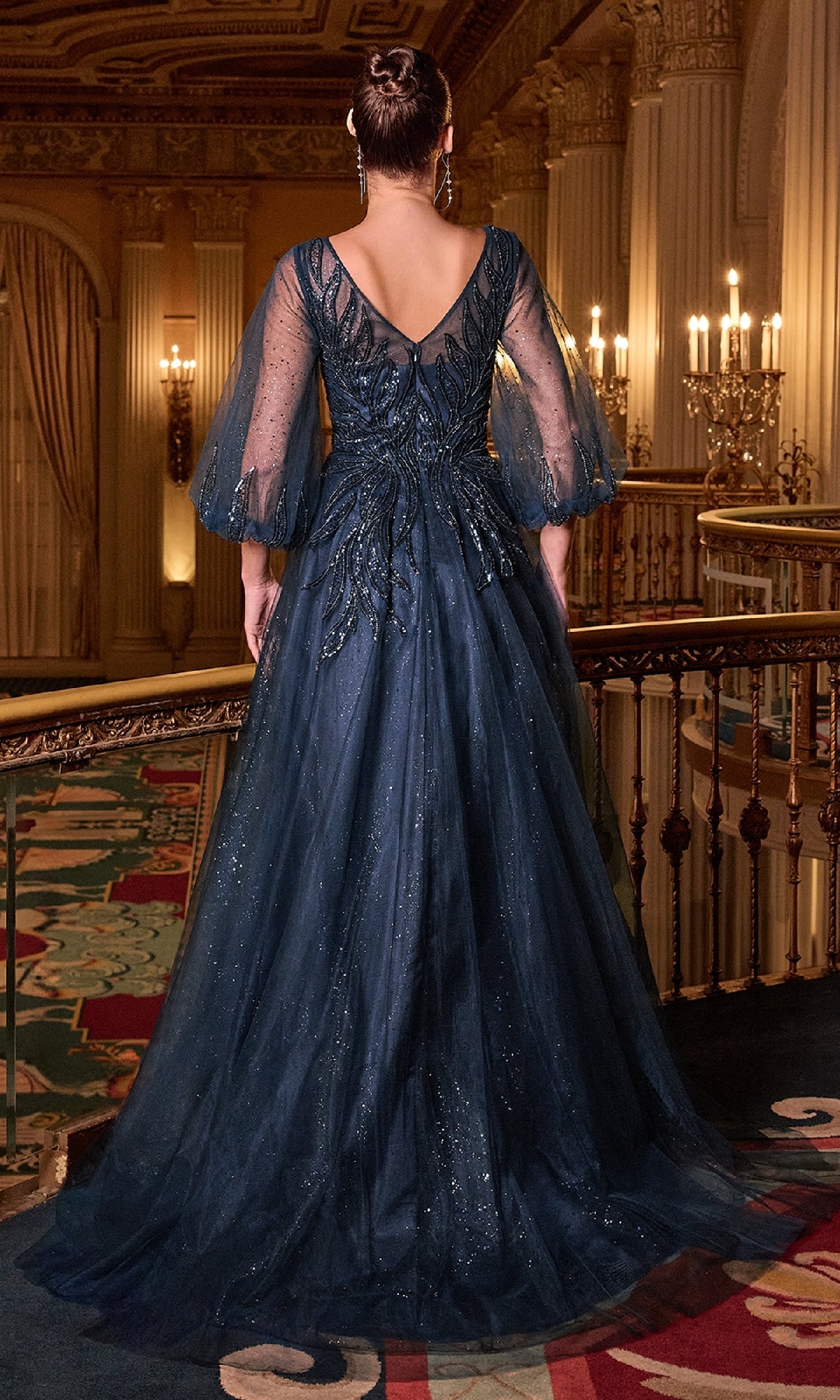 A woman in the Balloon-Sleeve Blue Formal Gown: Ladivine CD0101 stands with her back to the camera in a candlelit hall, the long navy blue dress with sheer balloon sleeves flowing elegantly to the floor.