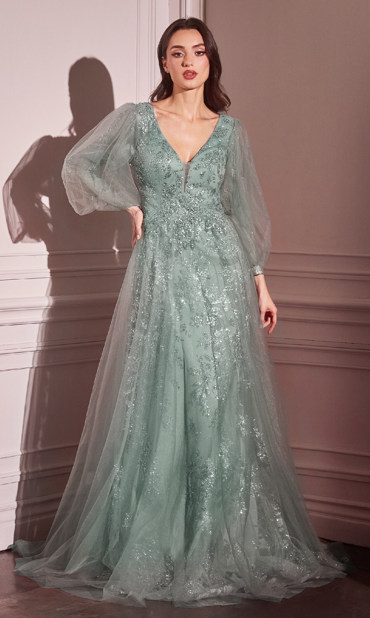 A woman wears the Ladivine CD0102 Sheer-Sleeve Long Prom Dress in light green, featuring a V-neck, sheer long sleeves, and floral sequin embellishments, standing before a white paneled wall.