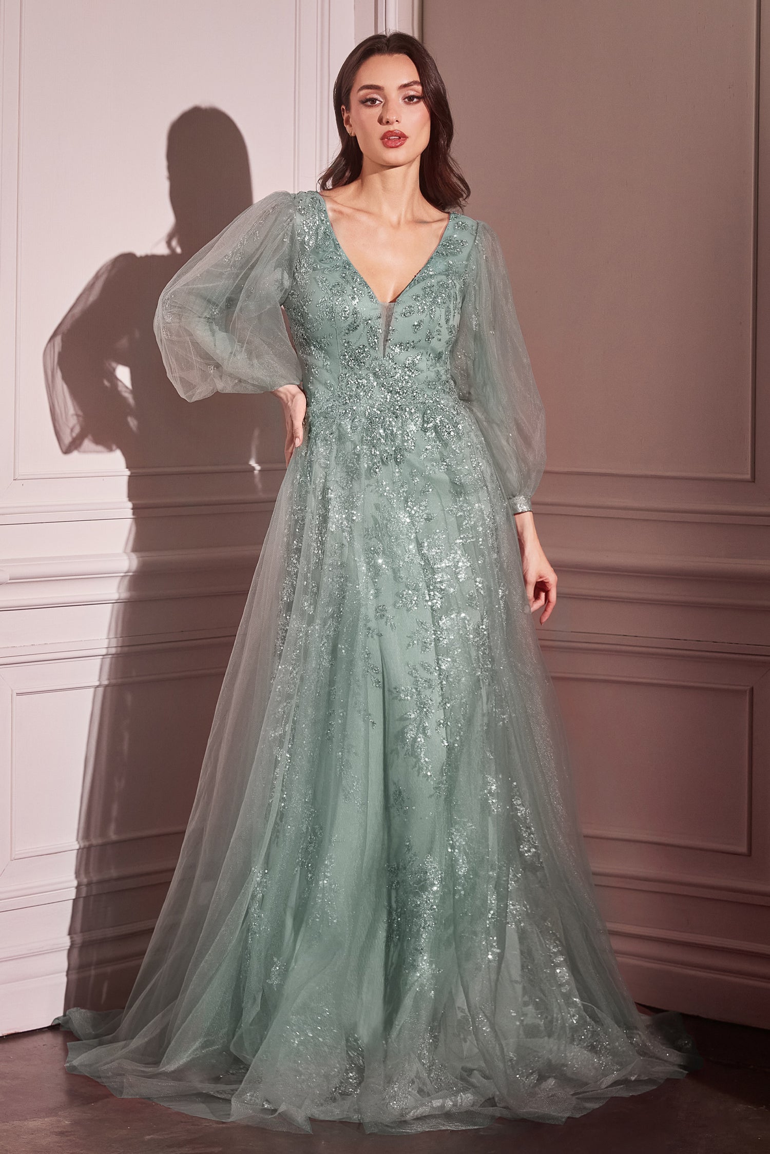 A woman stands indoors in the Ladivine CD0102 Sheer-Sleeve Long Prom Dress, a sage green, floor-length gown featuring sheer long sleeves, floral embellishments, and a deep V-neckline.
