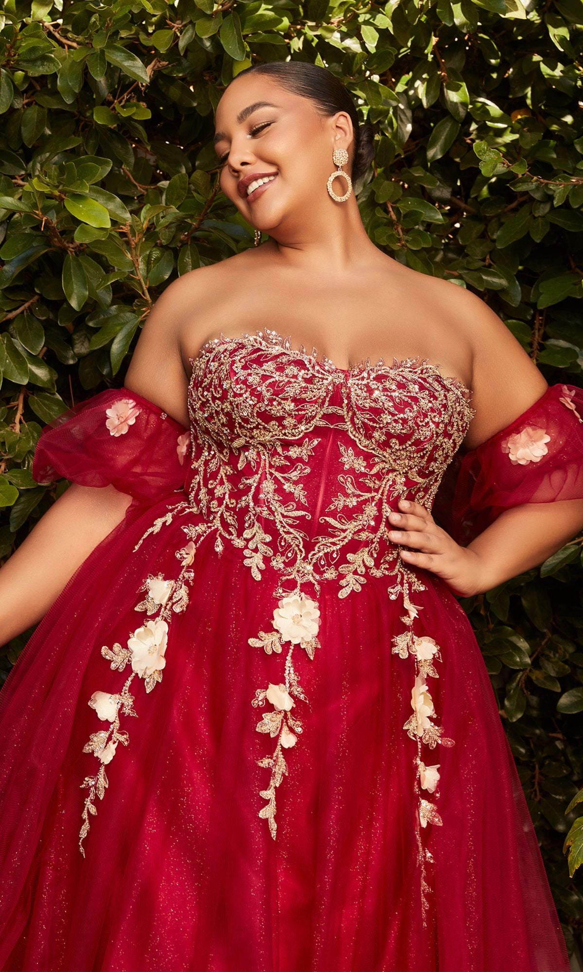 A woman smiles with closed eyes in front of green foliage, wearing the Plus-Size Strapless Prom Ball Gown CD0191C—an off-shoulder red dress with gold floral embroidery and a corset-style bodice.