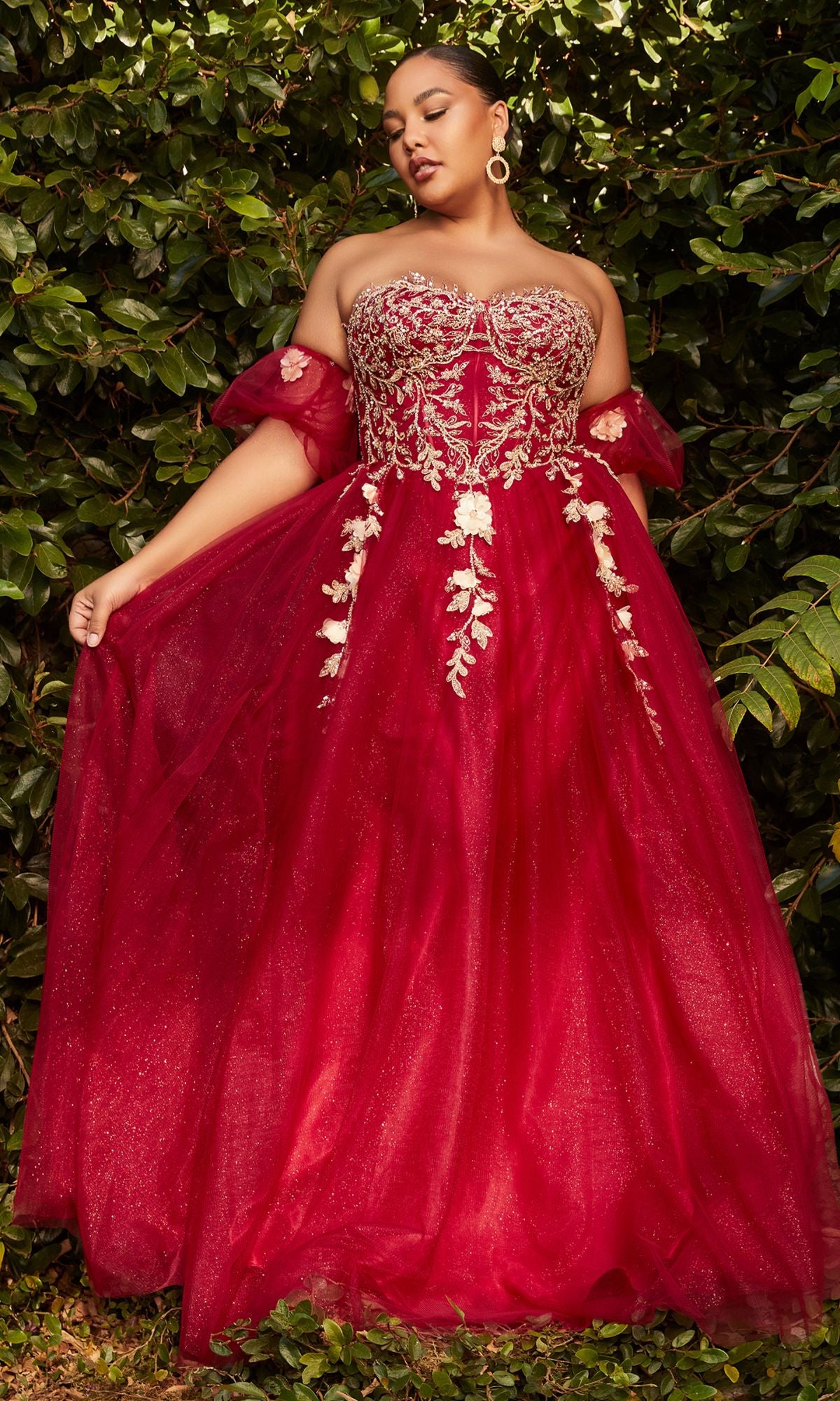 A woman stands before dense green foliage wearing the Plus-Size Strapless Prom Ball Gown CD0191C, featuring a corset-style bodice, gold embroidery, and red tulle fabric.