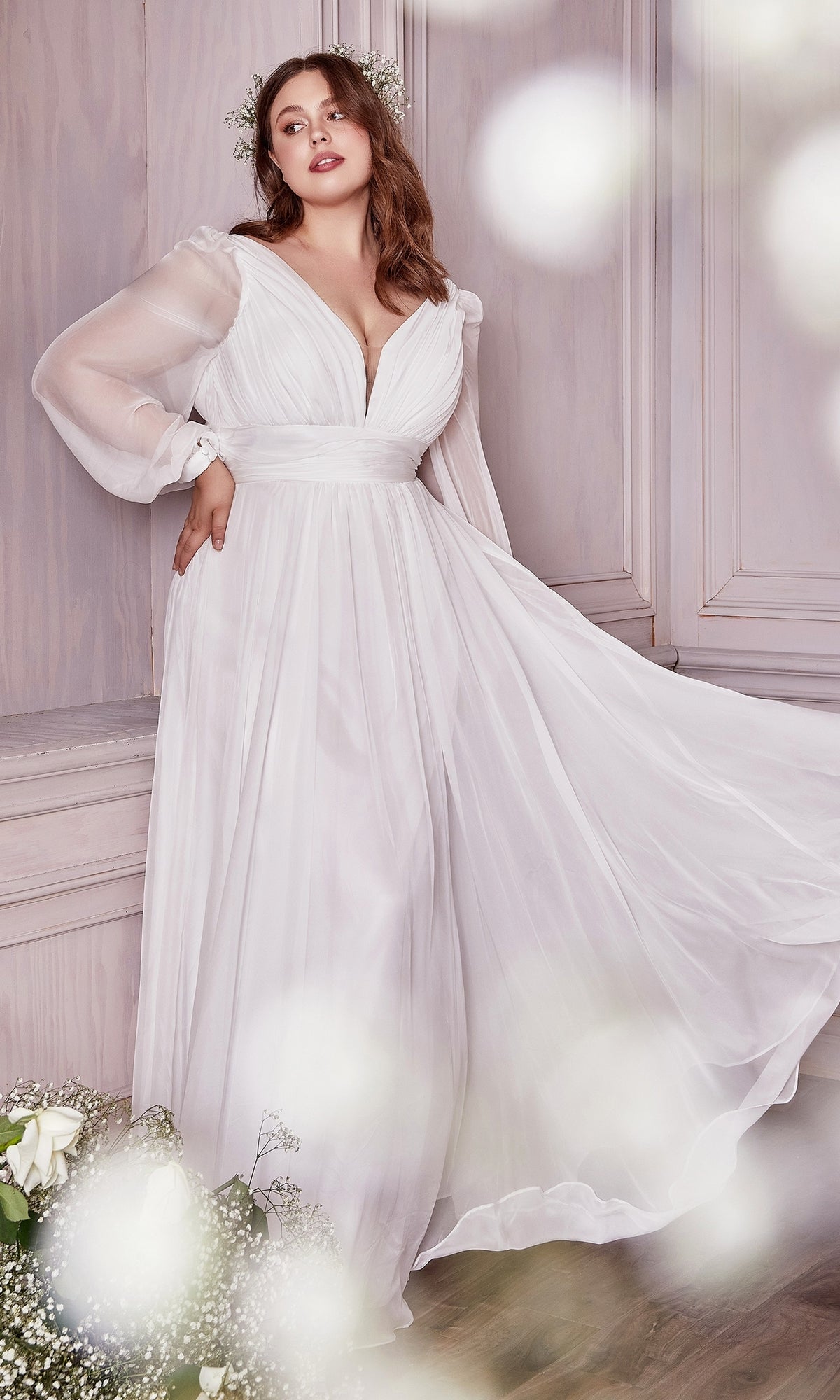 A woman in a long white chiffon formal dress with long sleeves stands indoors by a wall, surrounded by white flowers and soft lighting—an elegant and affordable option under $200.