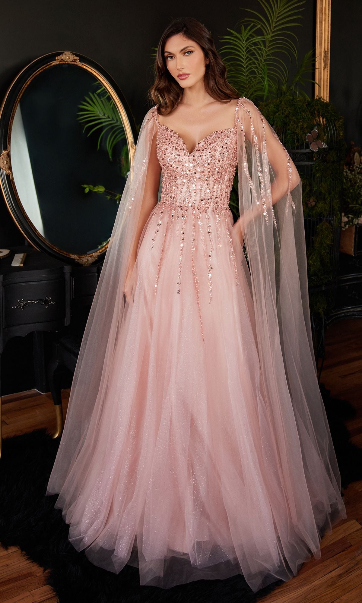 A woman stands indoors in the Cape-Sleeve Long Beaded Prom Dress CD0204, a pink sequined A-line gown with sheer cape sleeves, in front of a round mirror and dark furnishings.