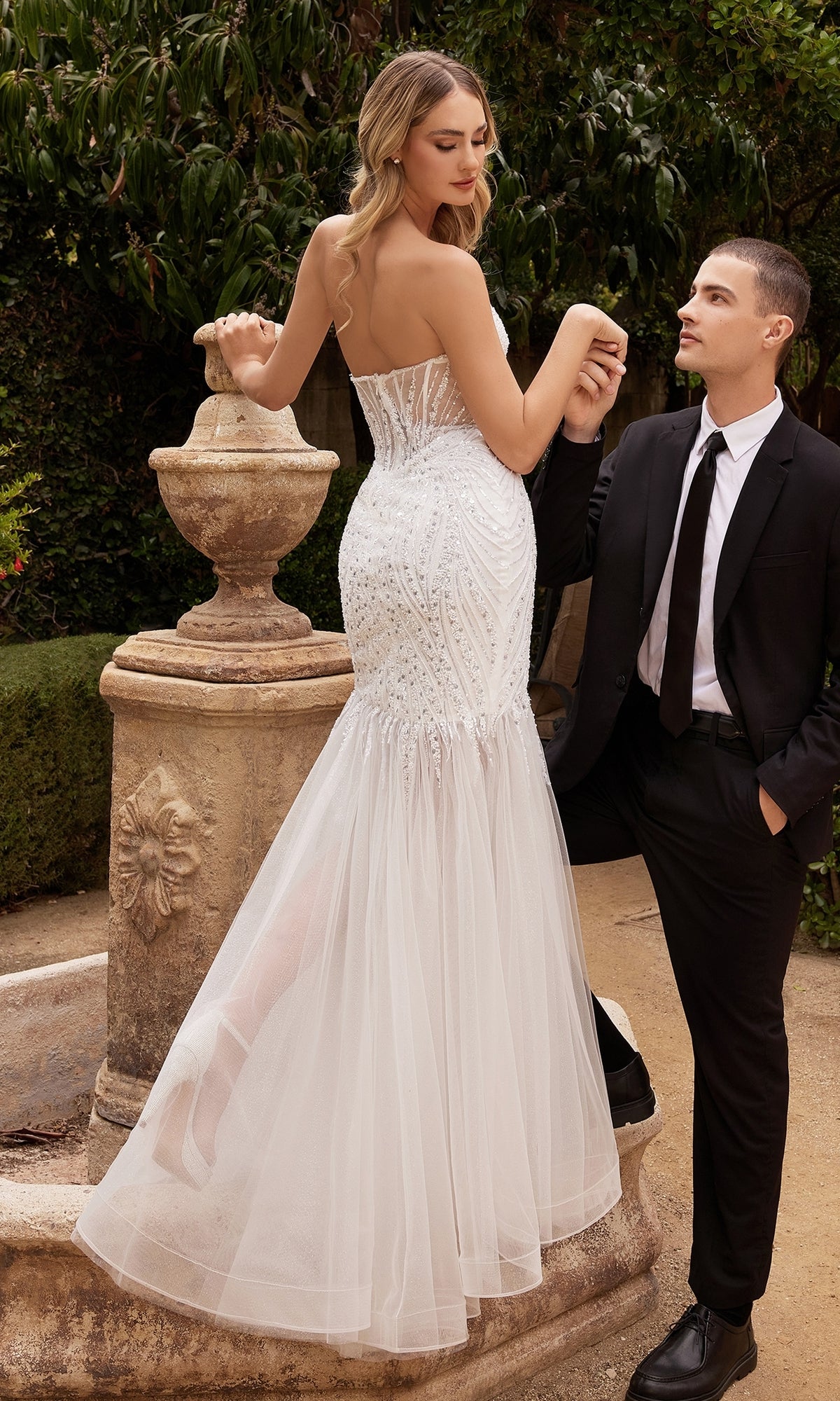 A woman wearing the Strapless Long White Mermaid Wedding Gown CD0215W stands by a stone fountain as a man in a black suit and tie holds her hand and looks at her.