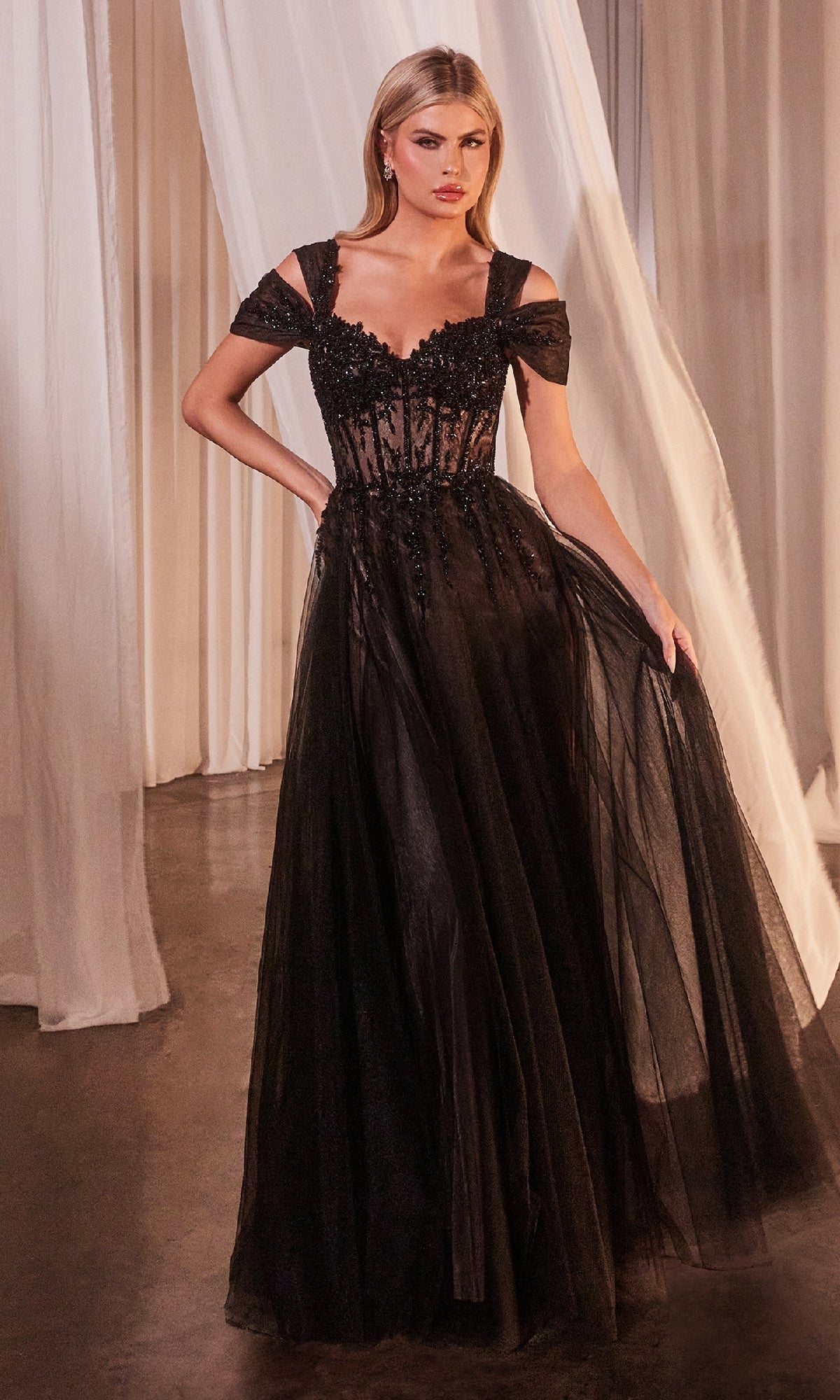 A woman stands indoors wearing the Lace-Up Long Black Prom Dress: Ladivine CD0237, an off-the-shoulder, floor-length A-line gown with sheer fabric and floral details, elegantly holding part of the skirt to showcase the formal look.