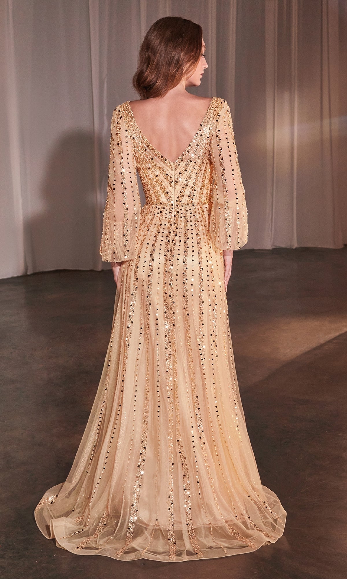 A woman indoors wears the Ladivine CD0240, a long sleeve beaded formal dress with a deep V-back and sheer sleeves. Her loose hair cascades down as she faces away, highlighting the elegance of this evening gown.