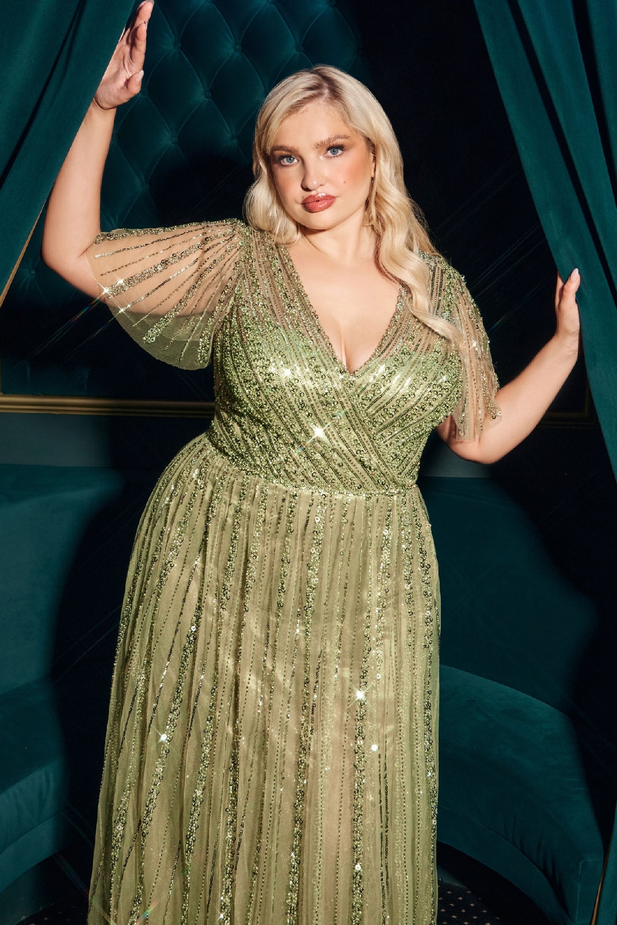 A woman with blonde hair poses between green velvet curtains in a dimly lit room, her Flutter-Sleeve Long Prom Dress: Ladivine CD0241 sparkling as the light catches its beaded, floor-length fabric.