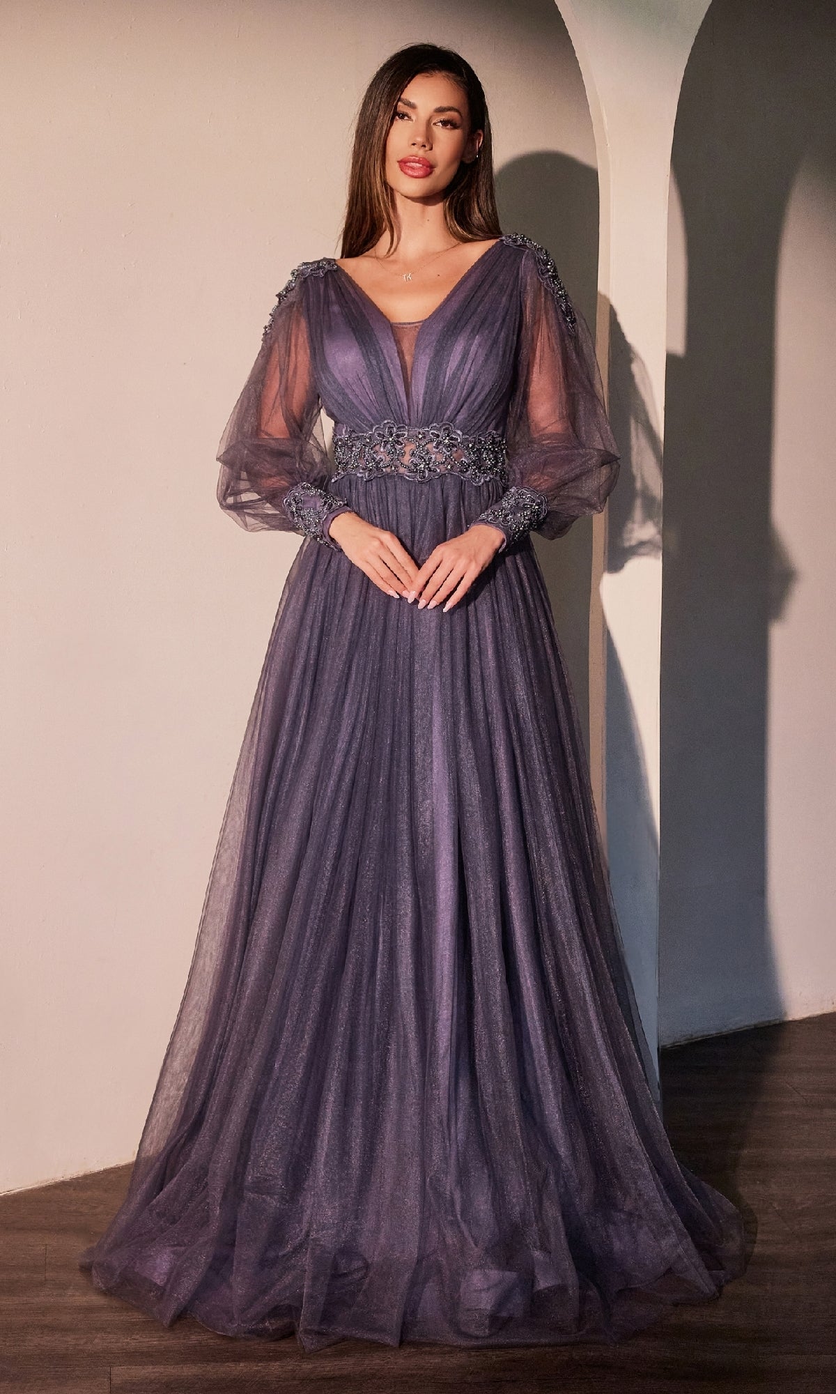 A woman stands indoors against a light wall and arched doorway, wearing the Ladivine CD0938 Long Sleeve Violet Formal Dress featuring sheer sleeves and floral waist embellishments.