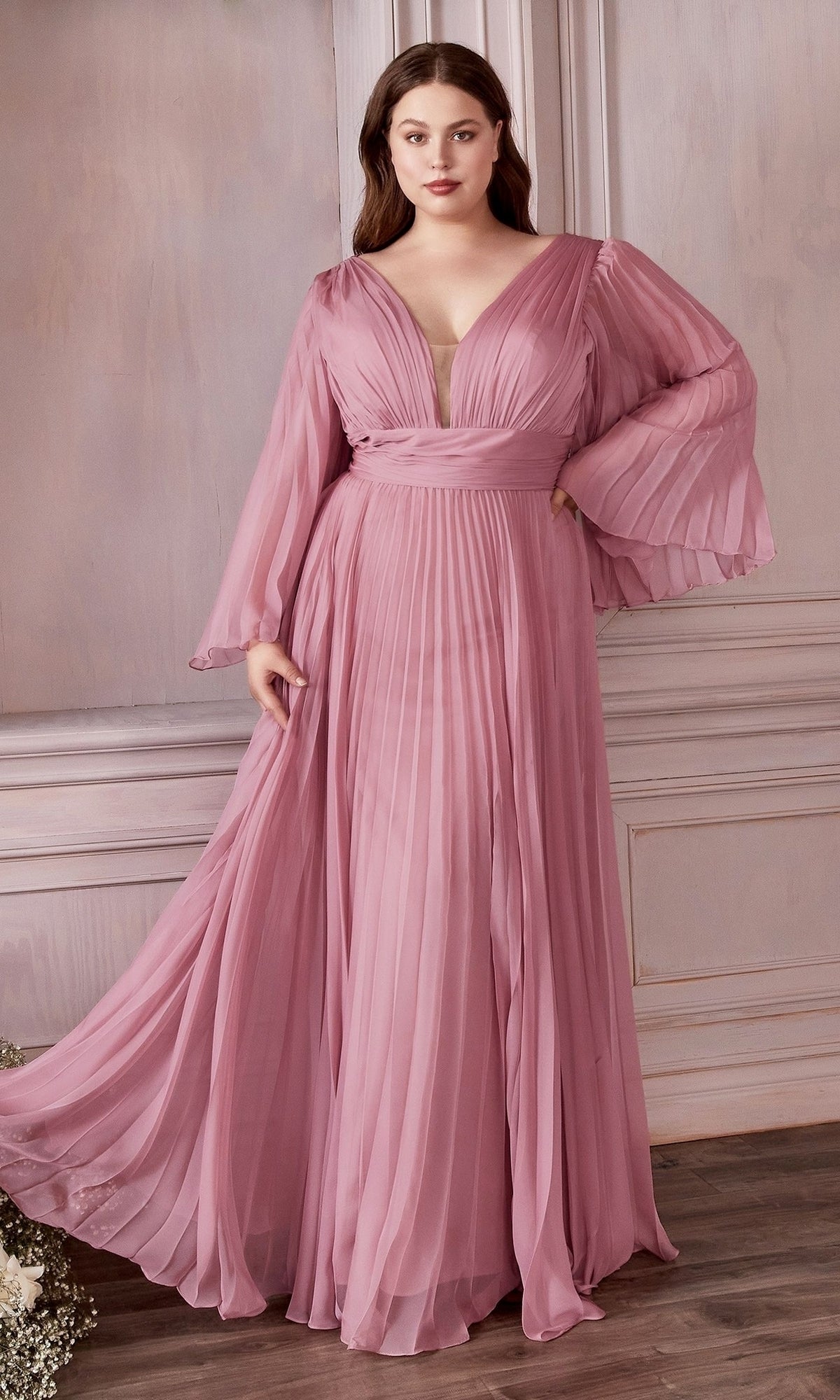 A woman stands indoors against a light ornate wall, wearing the Long Sleeve Plus-Size Long Formal Dress CD242C in blossom pink, which features sheer bell sleeves and elegant pleating.