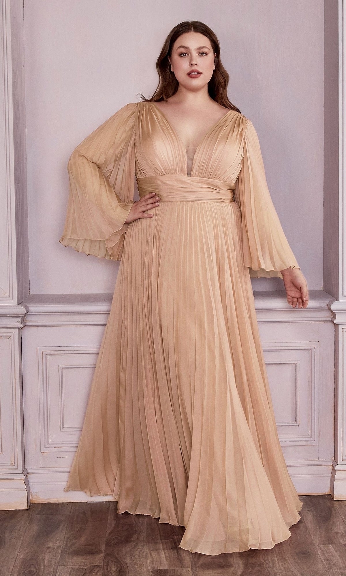 A woman stands indoors in the Long Sleeve Plus-Size Long Formal Dress CD242C, a champagne-colored, pleated gown with flared sleeves. Perfect for evening events, she is set against white paneled walls and a wooden floor.