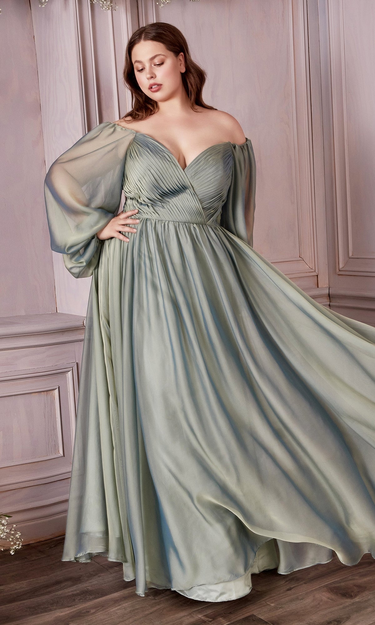 A woman stands in a softly lit, wood-paneled room wearing the Ladivine Plus-Size Off-Shoulder Prom Dress CD243C in sage green with sheer long sleeves.