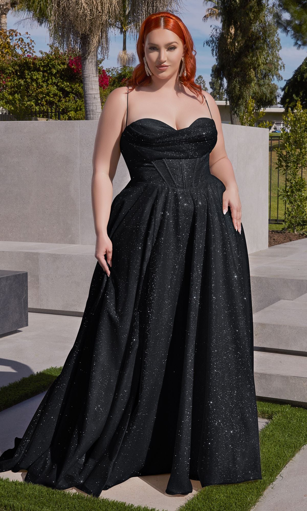 A red-haired woman stands outdoors on a paved area, wearing the Plus-Size Long Glitter Prom Dress CD252C, a black sparkly strapless gown, with greenery and concrete walls in the background.
