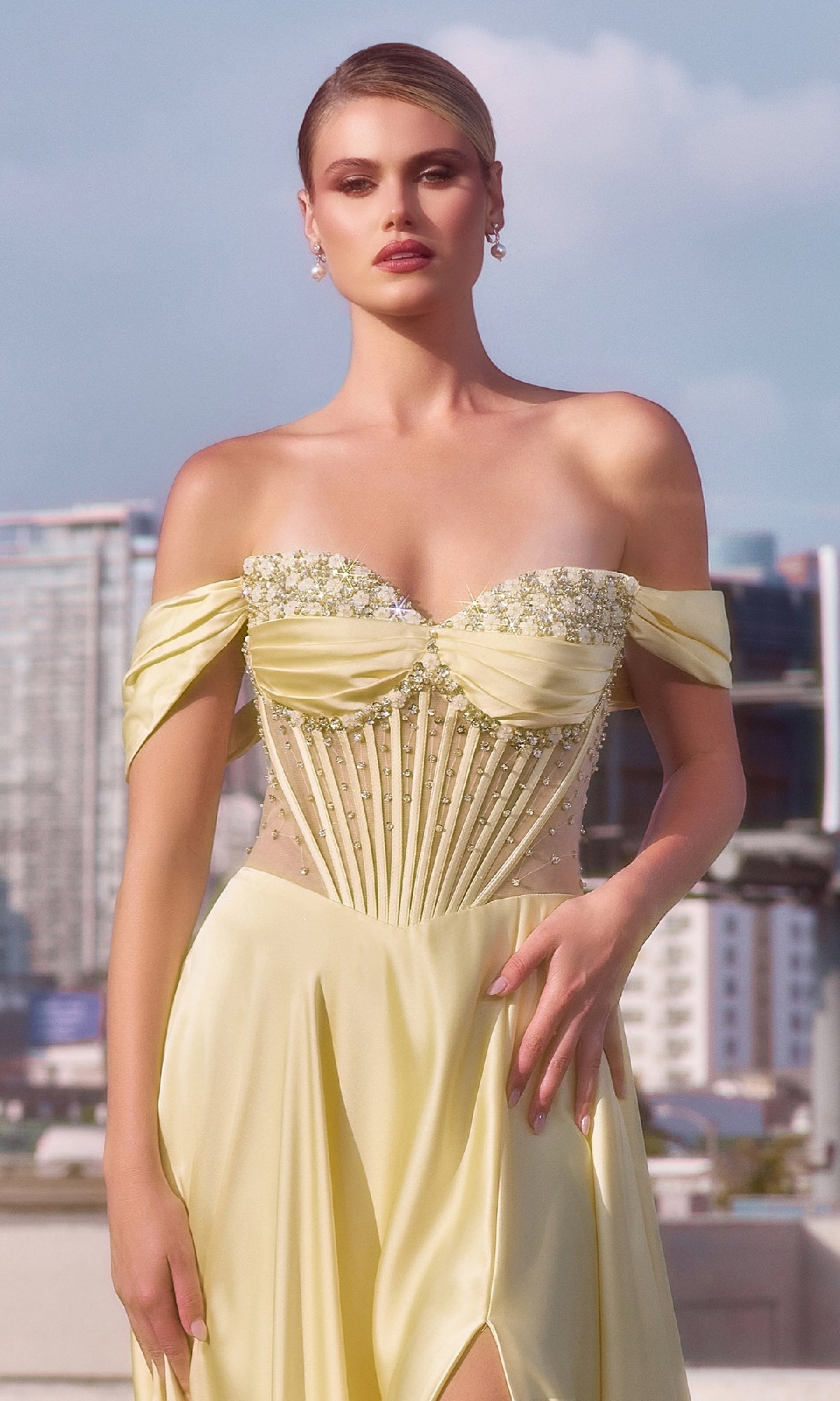 A woman in a yellow Ladivine CD297 strapless long A-line prom dress with beaded details stands outdoors before a city skyline.