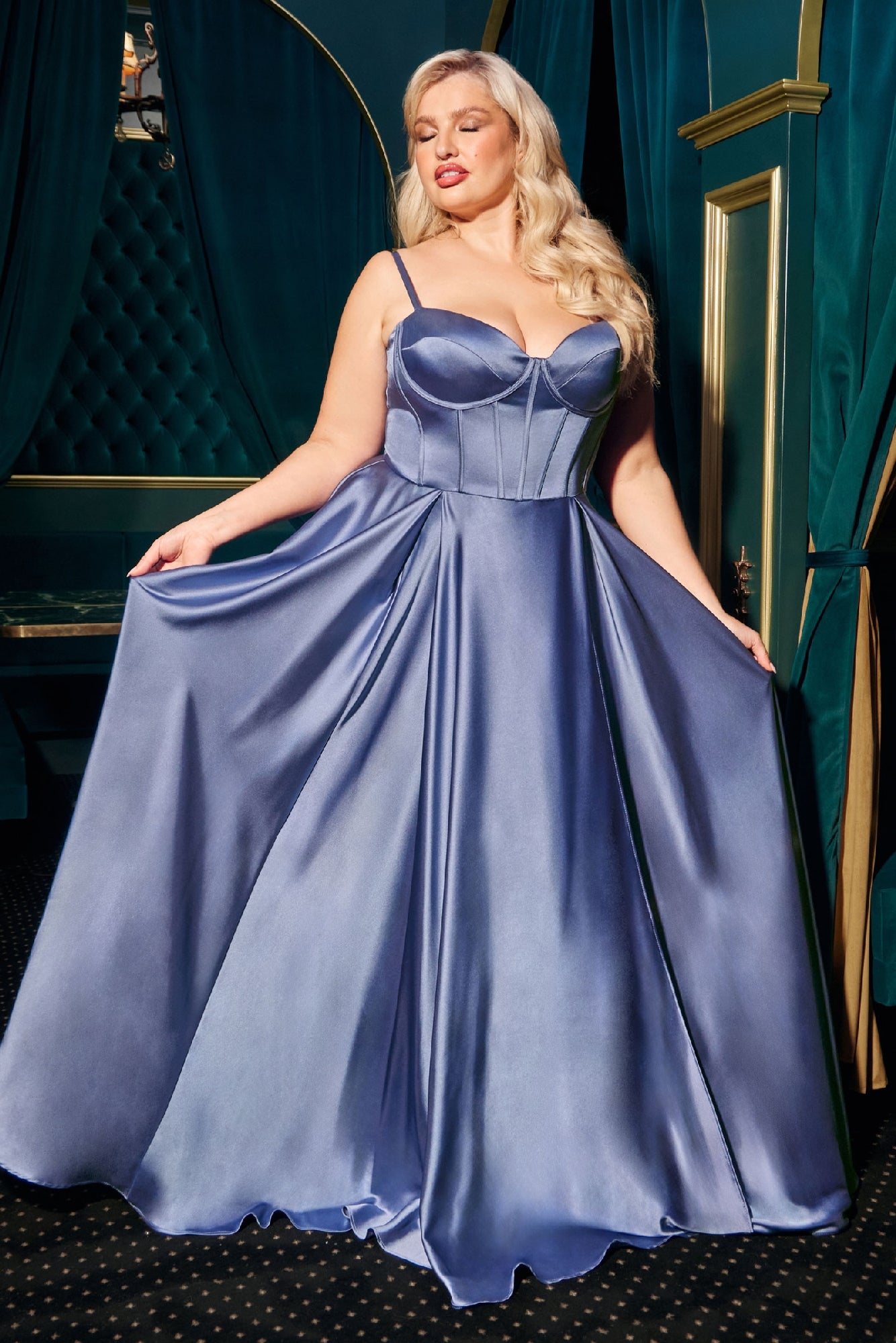 A woman in a shiny, floor-length Ladivine CD337C plus-size long A-line prom dress with a fitted bodice and thin straps holds out her gown in an elegant room with green and gold decor.