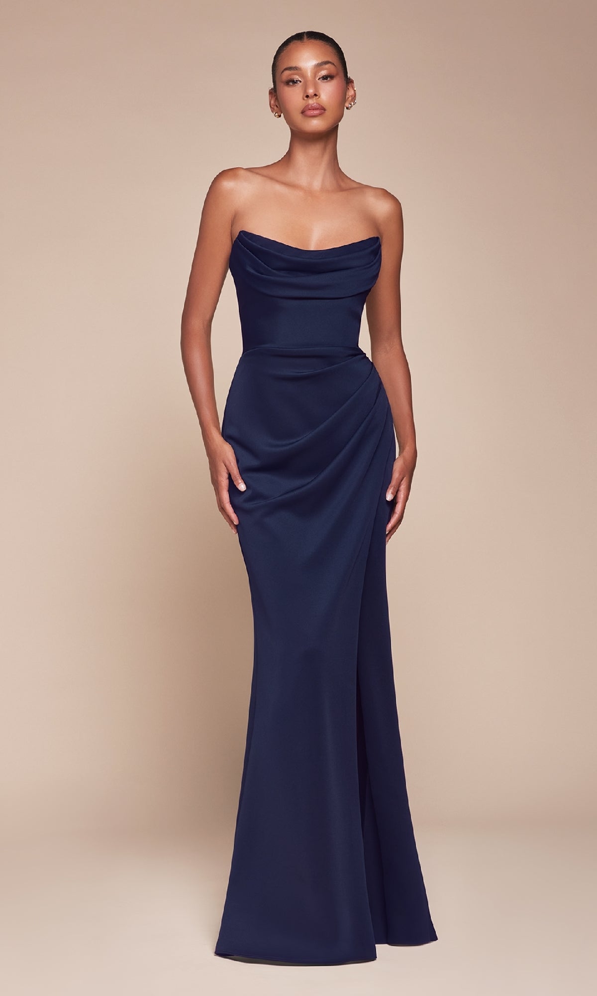 Wearing the Lace-Up Strapless Long Prom Gown: Ladivine CD355, this person stands against a plain beige background—an elegant floor-length navy blue dress with a draped bodice and side gathering, ideal for formal occasions.