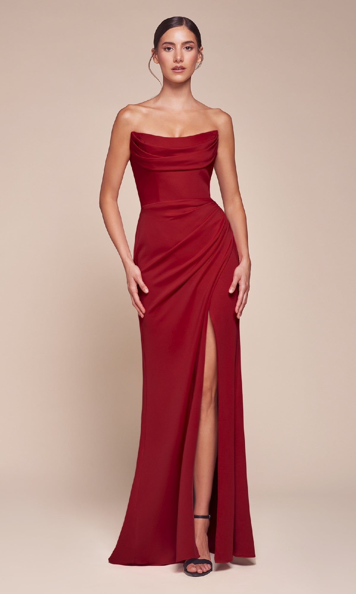 A woman wears the Lace-Up Strapless Long Prom Gown: Ladivine CD355 in vibrant red, with a draped bodice and high slit, standing against a plain beige background.