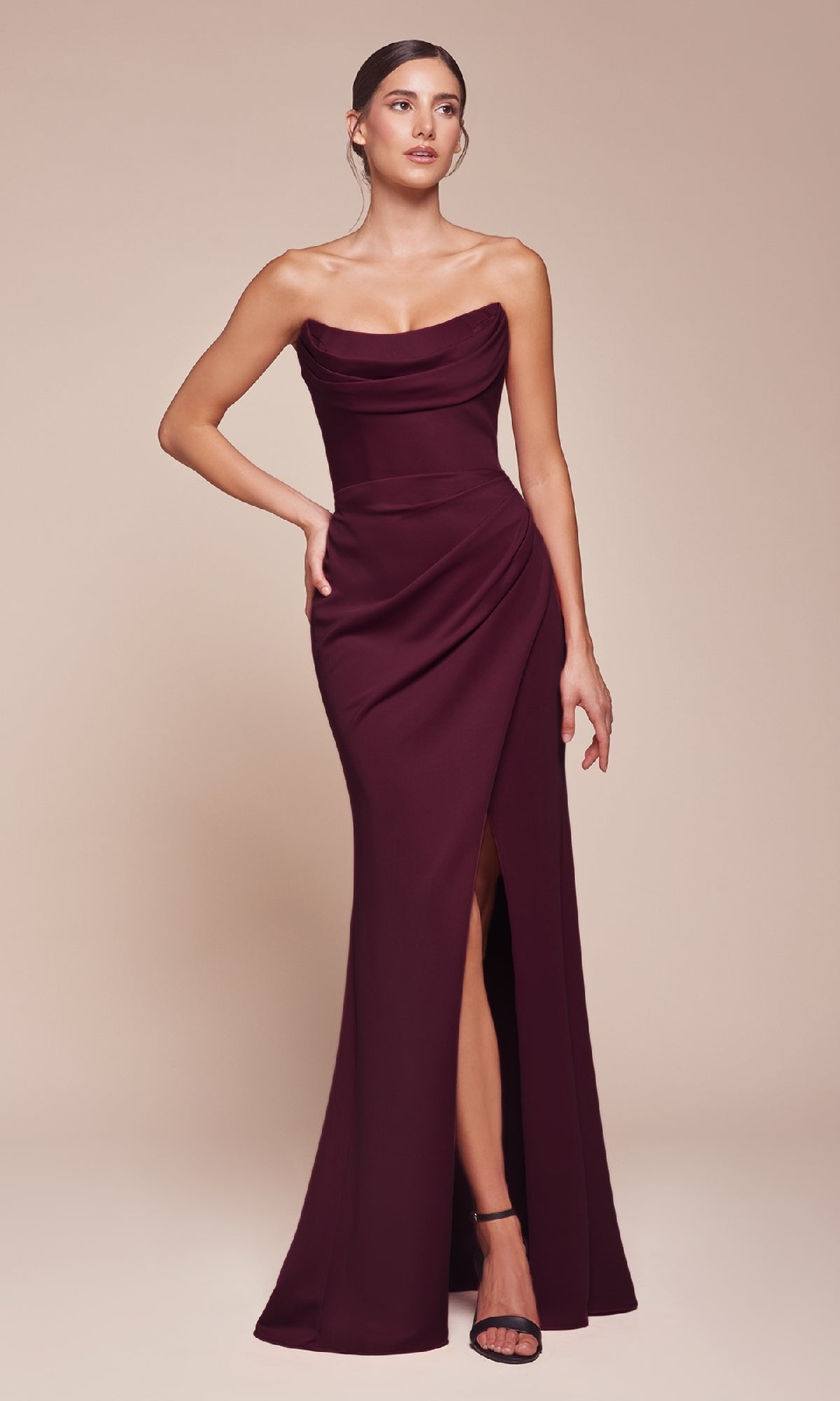 A woman in the Lace-Up Strapless Long Prom Gown: Ladivine CD355, a burgundy dress with a draped bodice and thigh-high slit, stands against a plain beige background.