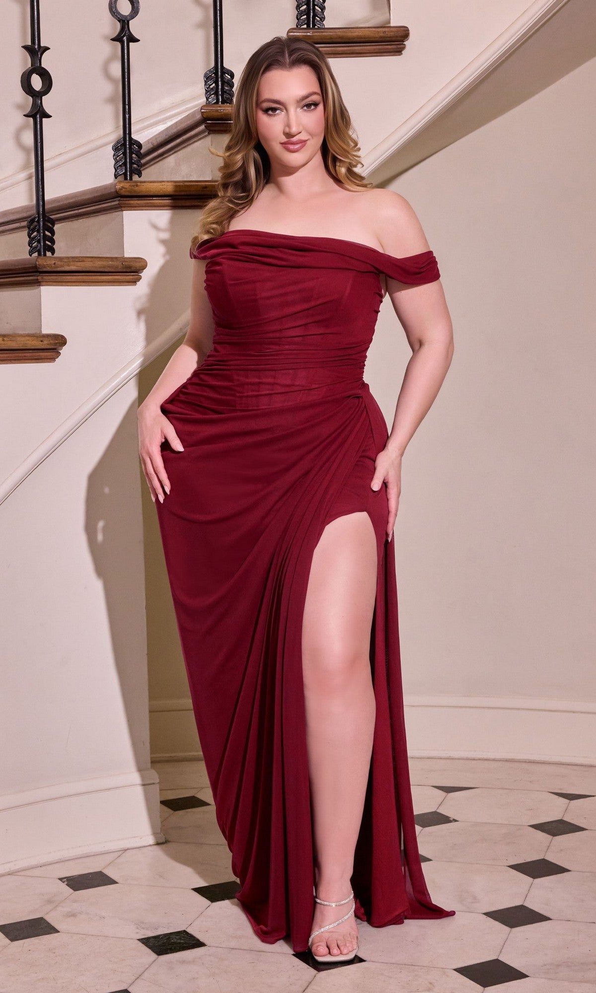 A woman wearing the Ladivine CD387C Long Plus-Size Prom Dress in burgundy, featuring an off-the-shoulder design, sheer corset bodice, and high slit, stands indoors before a staircase on a tiled floor.