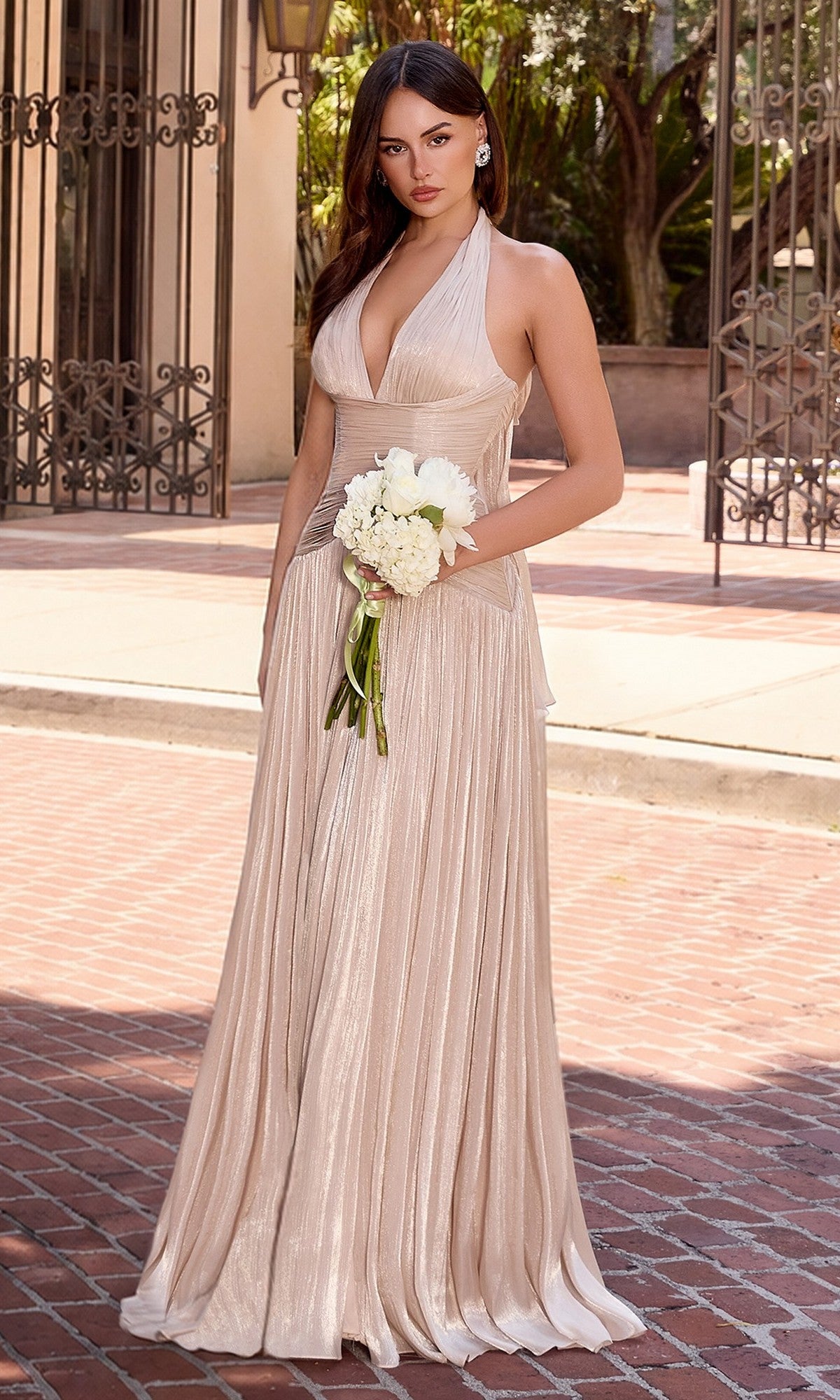 A woman stands on a brick walkway, holding a small white bouquet and wearing the Ladivine CD412 Long Ruched Halter Prom Dress in champagne gold, with buildings and greenery in the background.