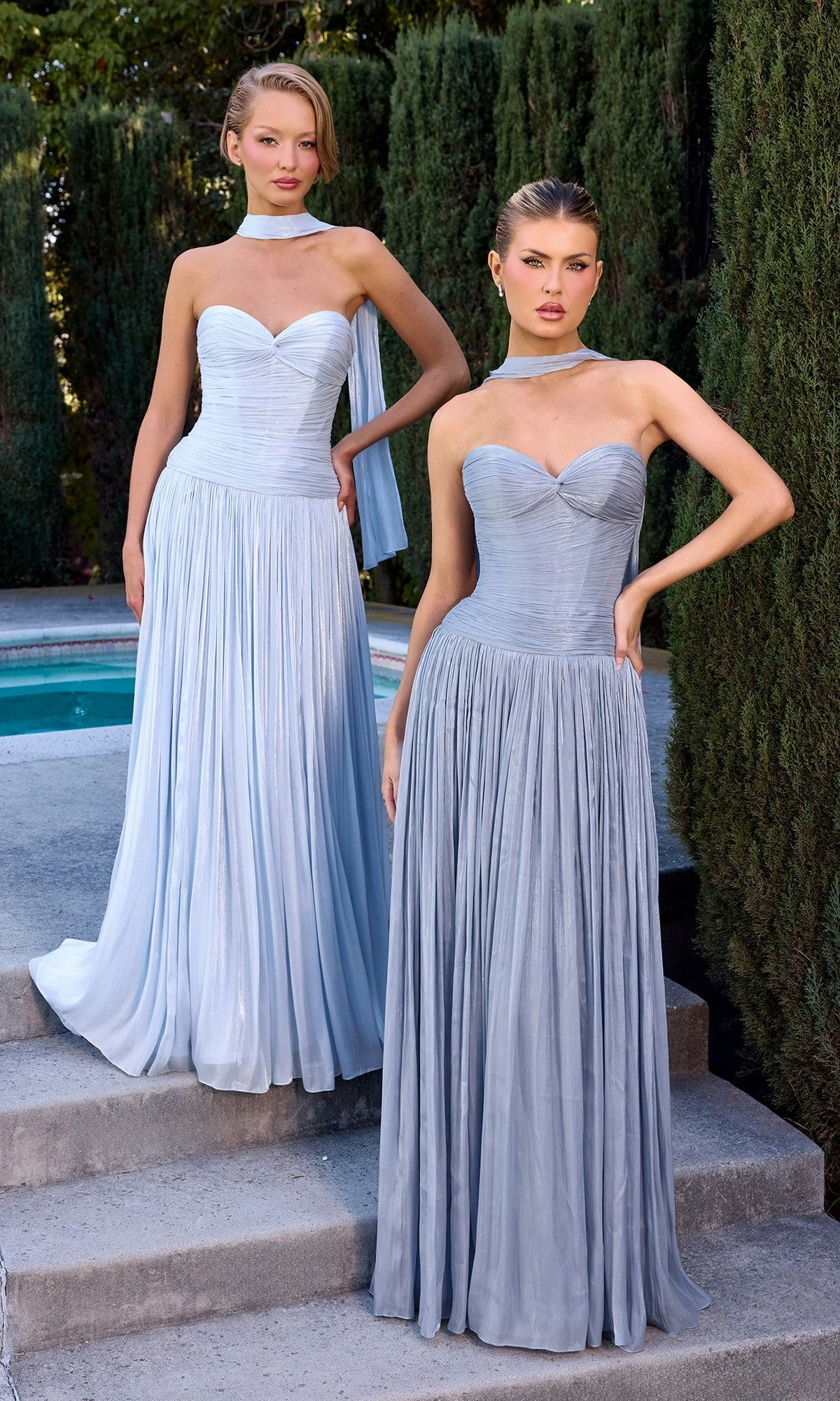 Two women stand outdoors by a pool, both wearing the Ladivine CD414 long prom dress in light blue chiffon with a-line skirts and matching neck scarves, trees visible in the background.