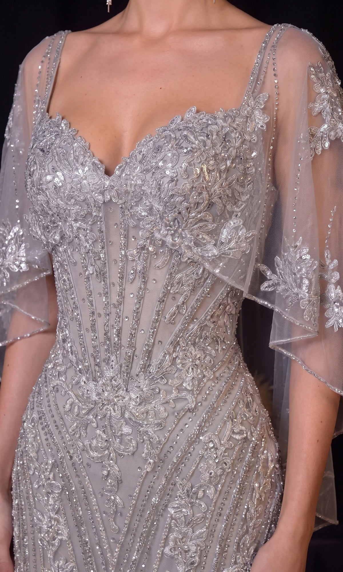 A person wearing the Ladivine CD637, a long silver beaded prom dress featuring a sheer corset bodice and sheer sleeves adorned with delicate floral patterns.
