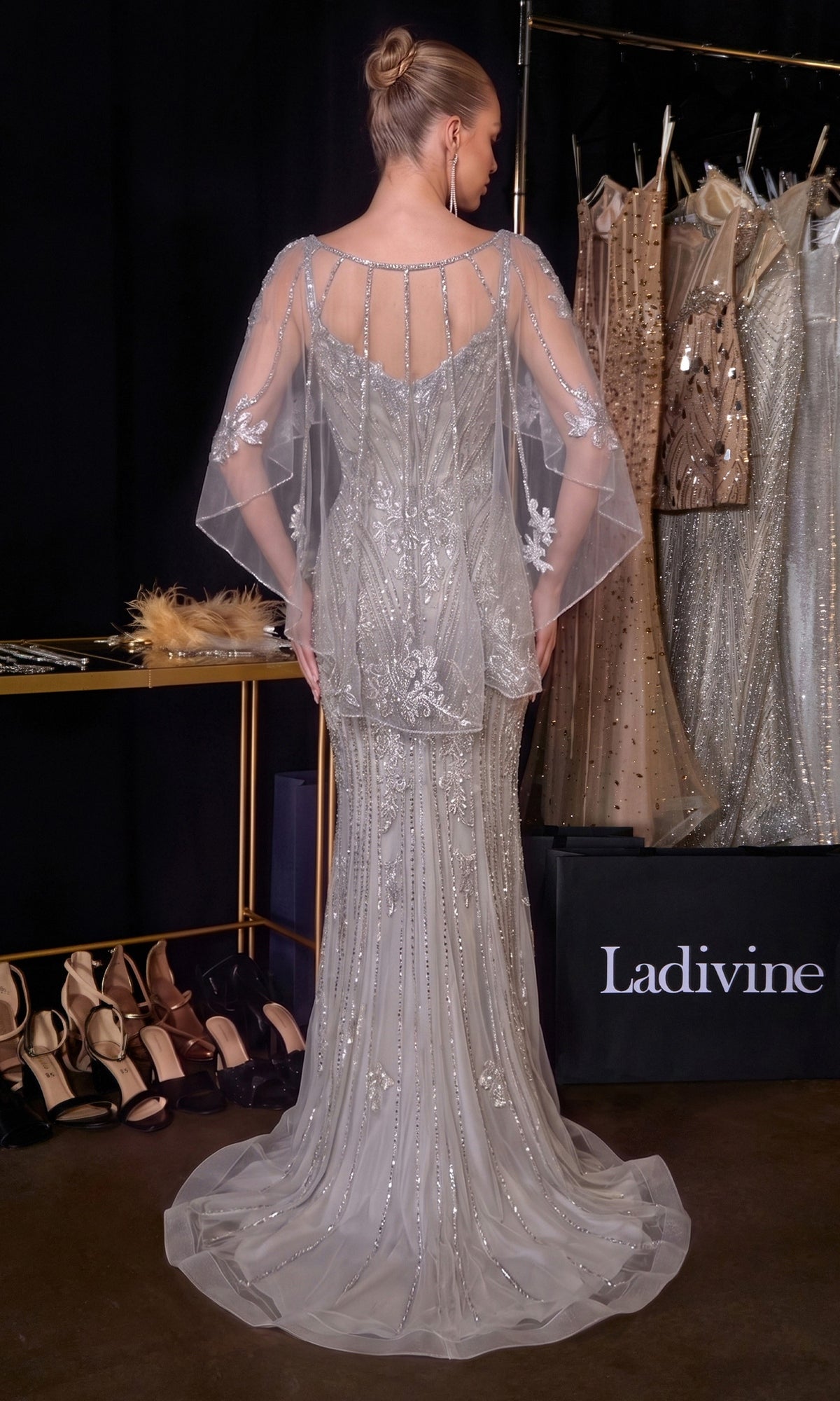 A woman wearing the Ladivine CD637 Long Prom Dress, featuring silver beading and a sheer cape, stands with her back to the camera near a rack of dresses and a shelf of high-heeled shoes.