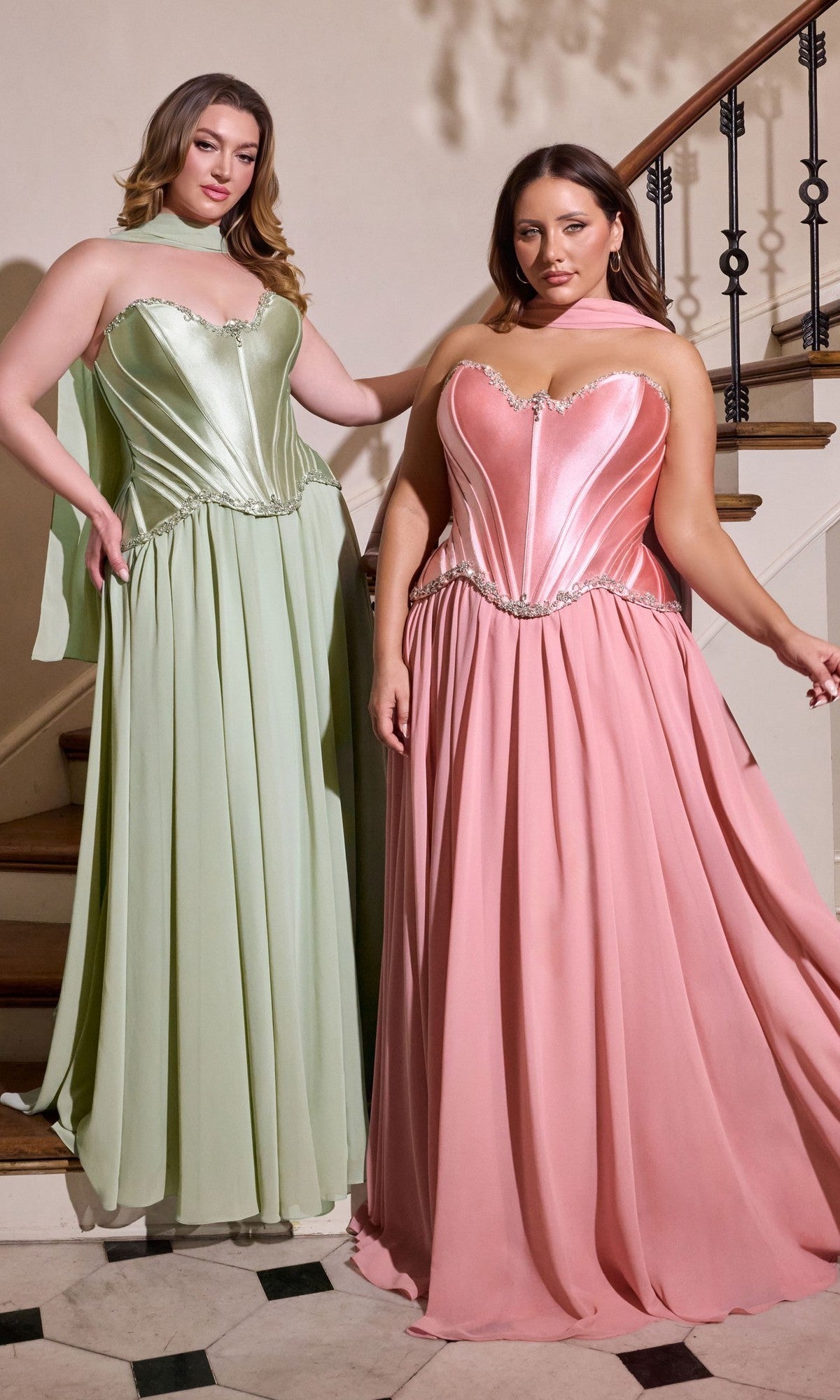 Two women stand together on a staircase indoors, one wearing the Long Plus-Size Prom Dress Ladivine CD668C in sage—a floor-length gown with silver embellishments—while the other wears a pink strapless gown with a corset bodice.