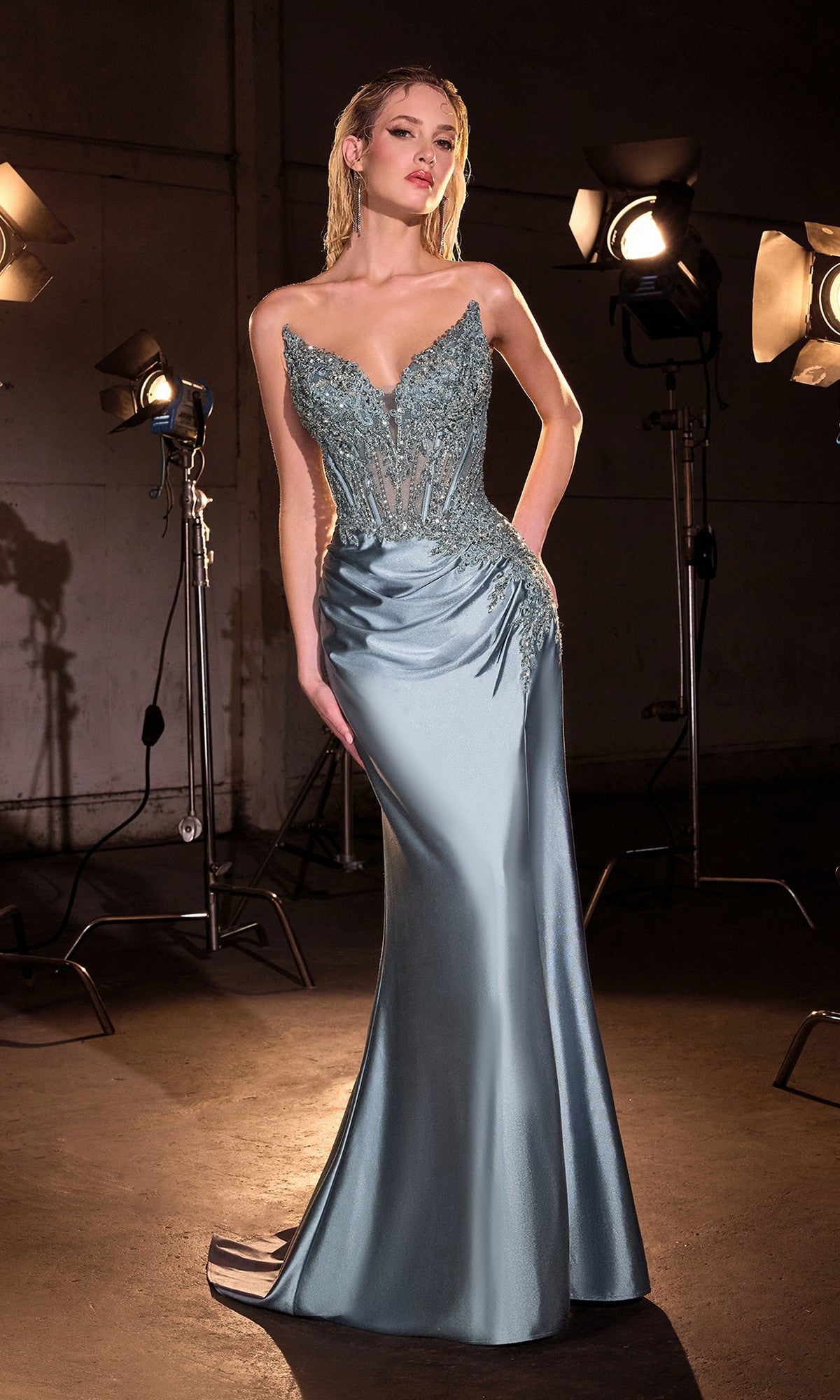 A woman stands in a studio wearing the Beaded-Corset Strapless Prom Gown: Ladivine CD714, a fitted light blue satin dress with beaded details, illuminated by studio lights.