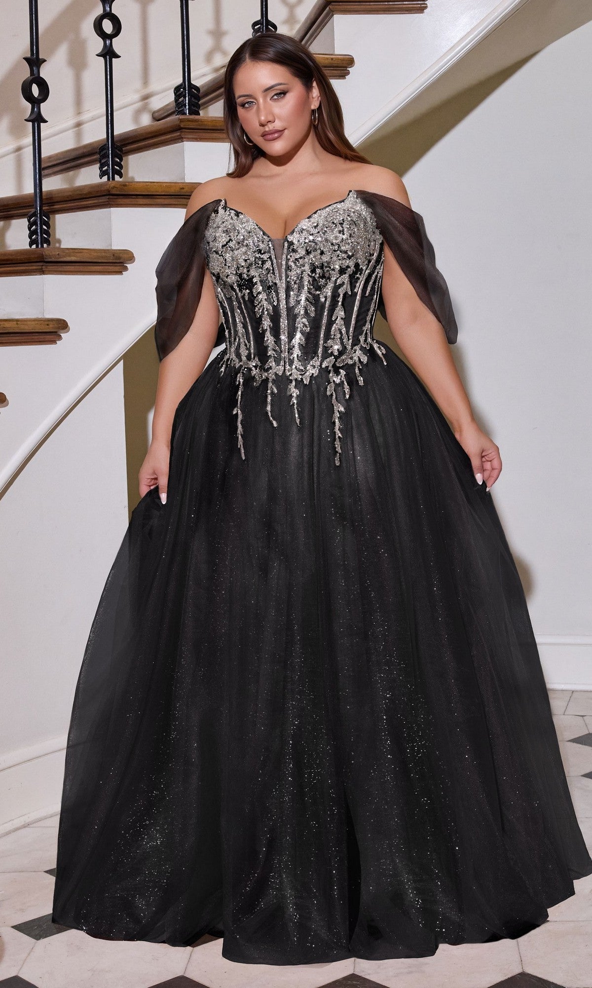 A woman stands on a marble floor near a staircase, wearing the Long Plus-Size Prom Dress Ladivine CD721C in Black/Silver, featuring an off-the-shoulder glitter tulle ball gown with an embellished corset bodice and full skirt.