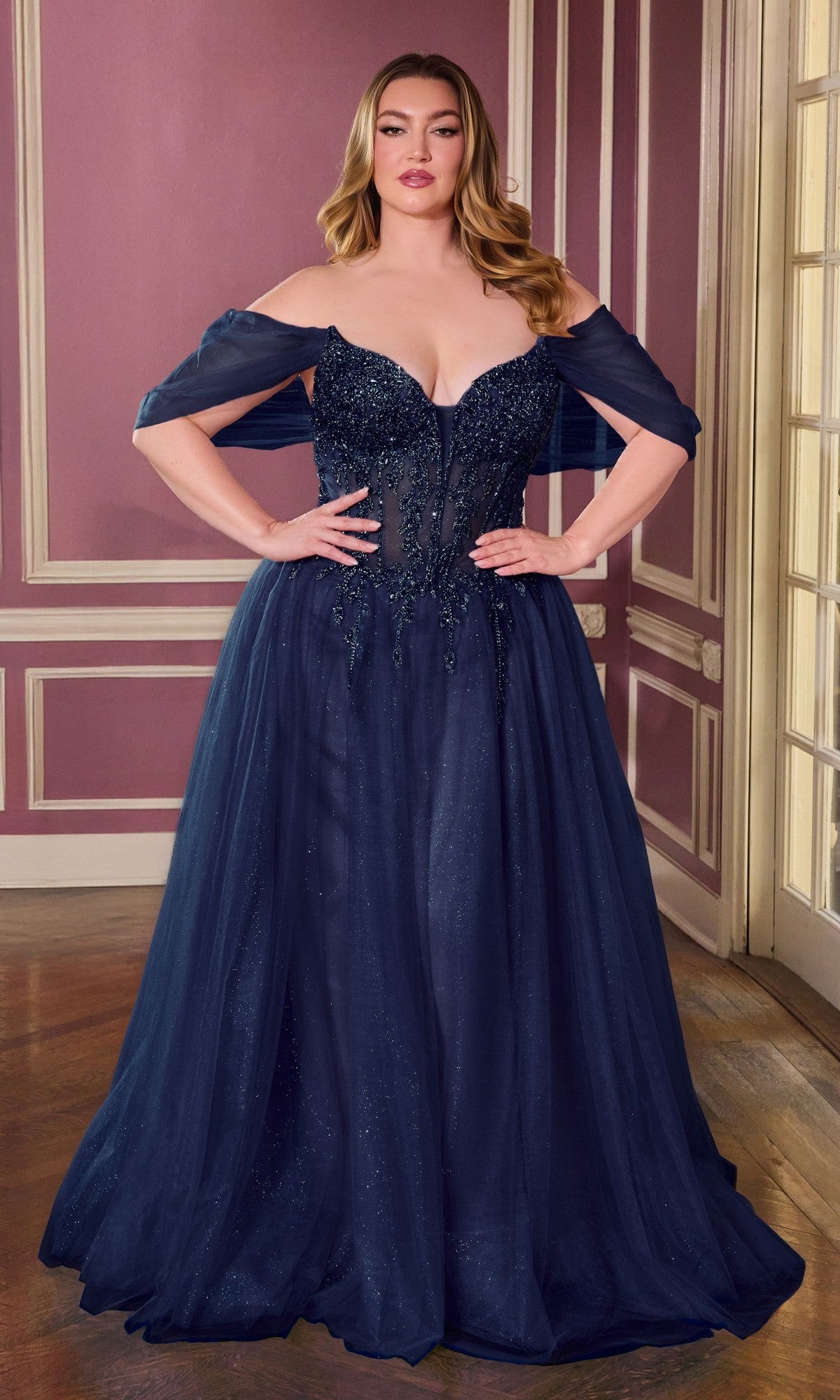 A woman stands indoors in the Ladivine CD721C, a long plus-size navy prom dress with an embellished corset bodice and off-the-shoulder style—an elegant choice for formal events.