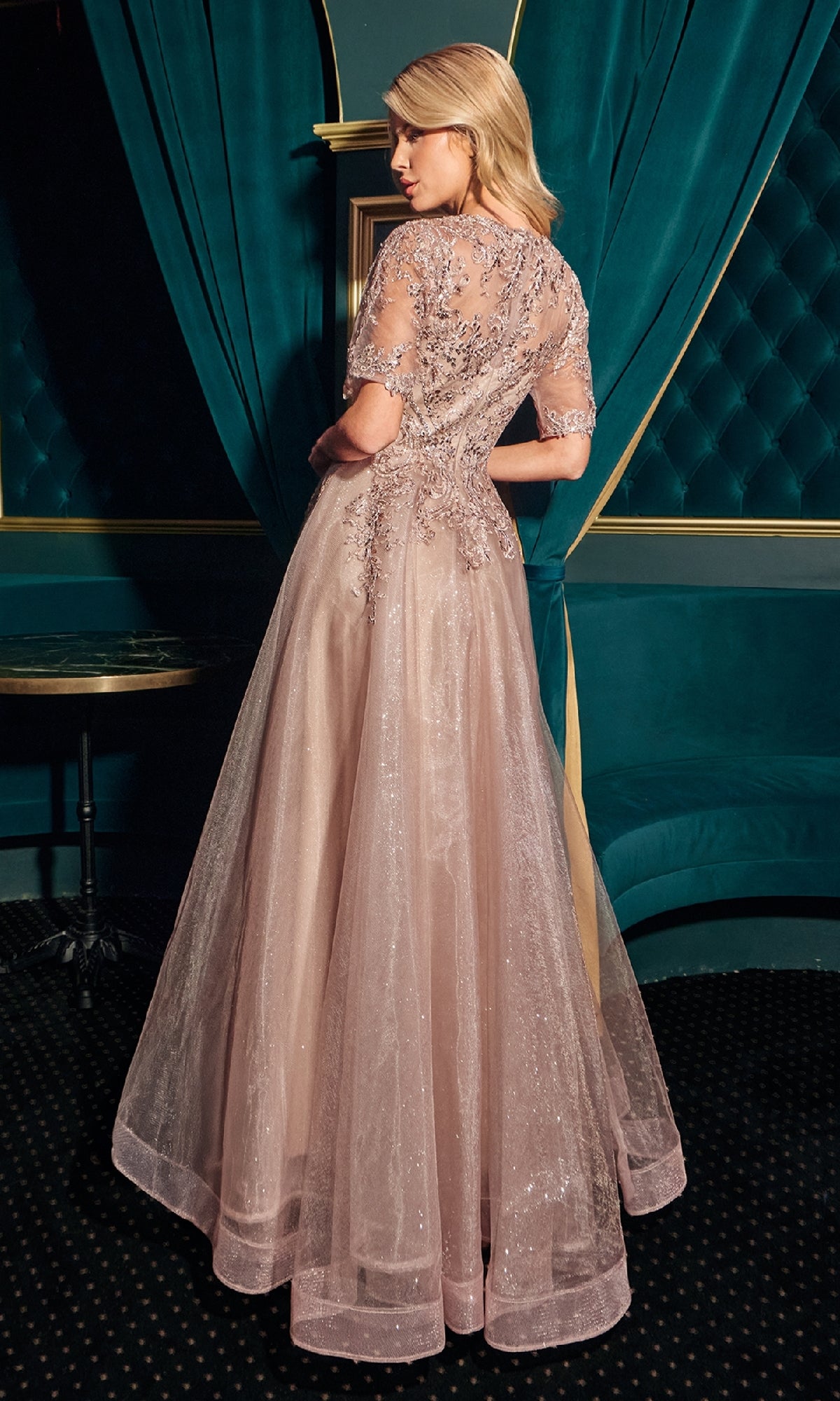 A woman in a blush pink, floor-length Ladivine CD726 short-sleeve glitter formal dress with sheer overlay stands with her back to the camera in an elegant, teal-accented room.