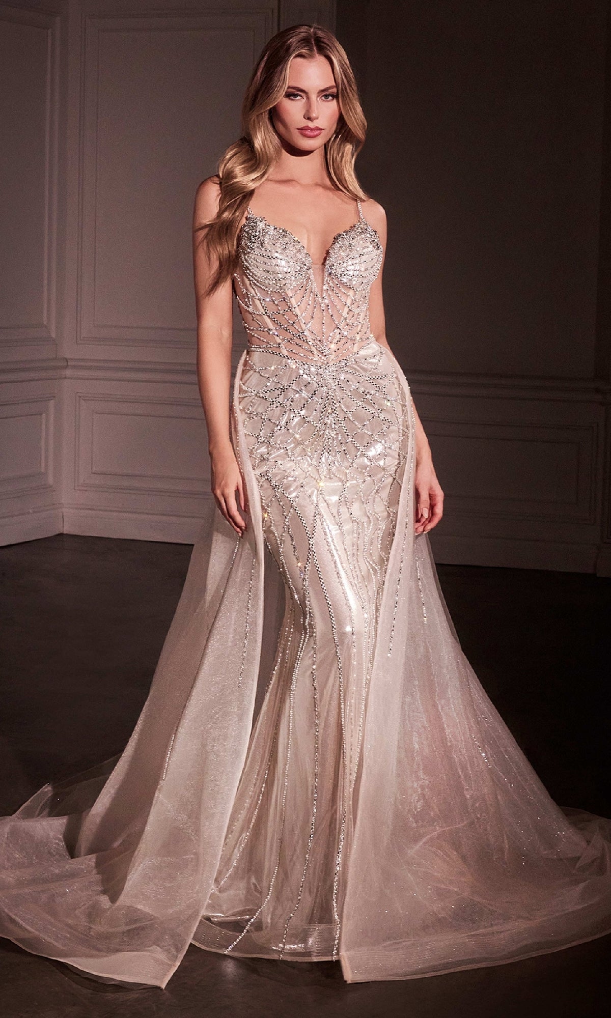 A woman stands in a softly lit room wearing the Ladivine CD763—an Overskirt Long Glitter Prom Dress with a fitted, sheer design, silver embellishments, and a flowing removable overskirt.