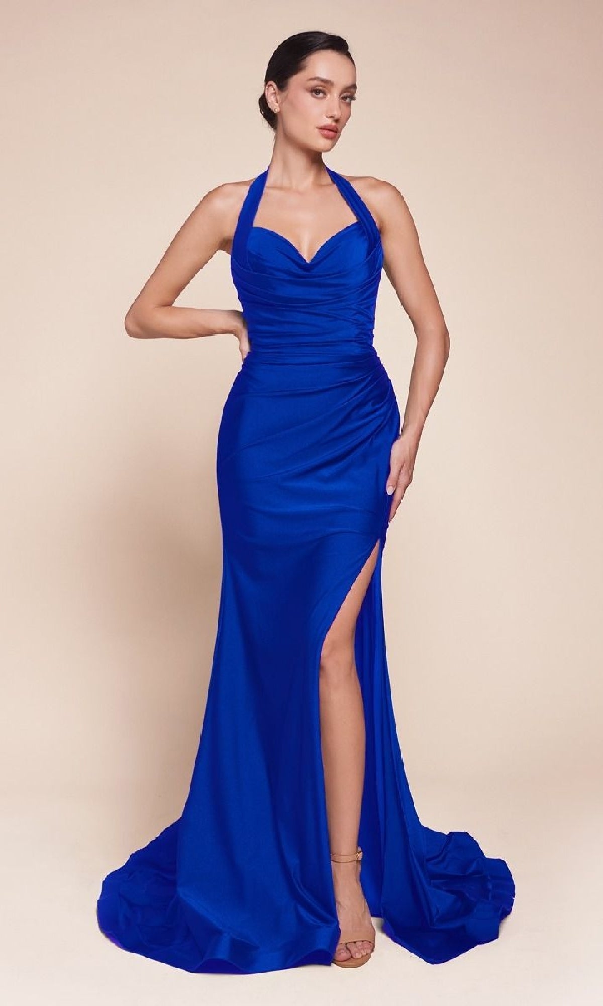 A woman in a royal blue Classic Long Halter Prom Dress: CLCD796 poses against a plain beige background, showcasing its elegant silhouette and thigh-high slit.