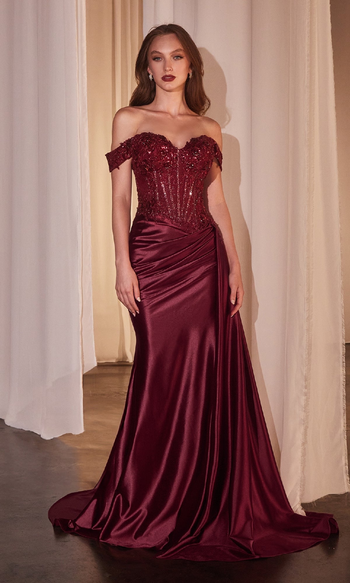 A woman stands indoors wearing the Off-the-Shoulder Long Prom Dress: Ladivine CD803—a floor-length burgundy gown with a fitted, sequined bodice and a satin draped skirt.