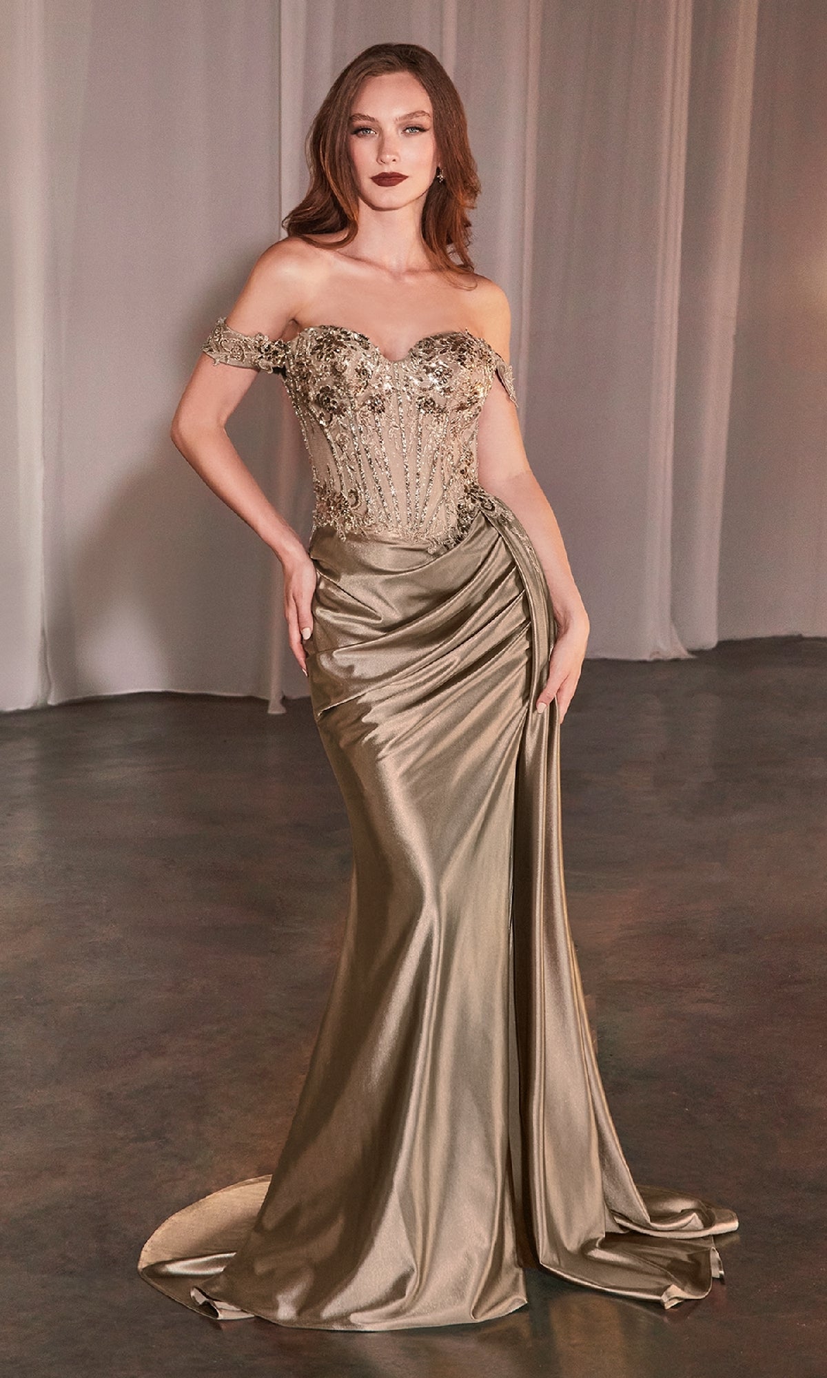A woman in the Ladivine CD803 Off-the-Shoulder Long Prom Dress, featuring a metallic gold beaded corset-style bodice, draped satin skirt, and slight train, stands on a dark floor with sheer curtains behind her.