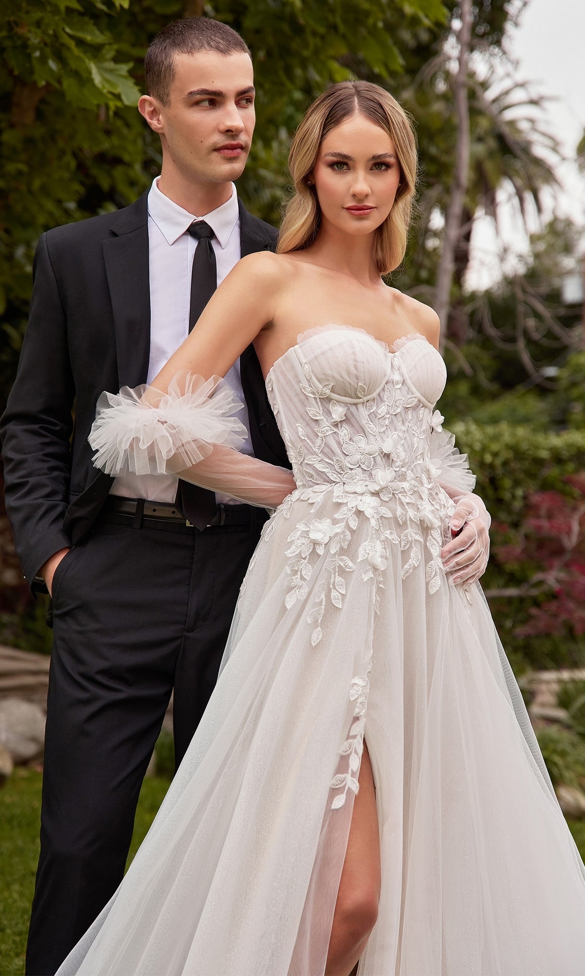 A man in a black suit poses outdoors with a woman wearing the Ladivine White Wedding Dress CD859W, an off-white strapless gown with delicate lace embroidery.
