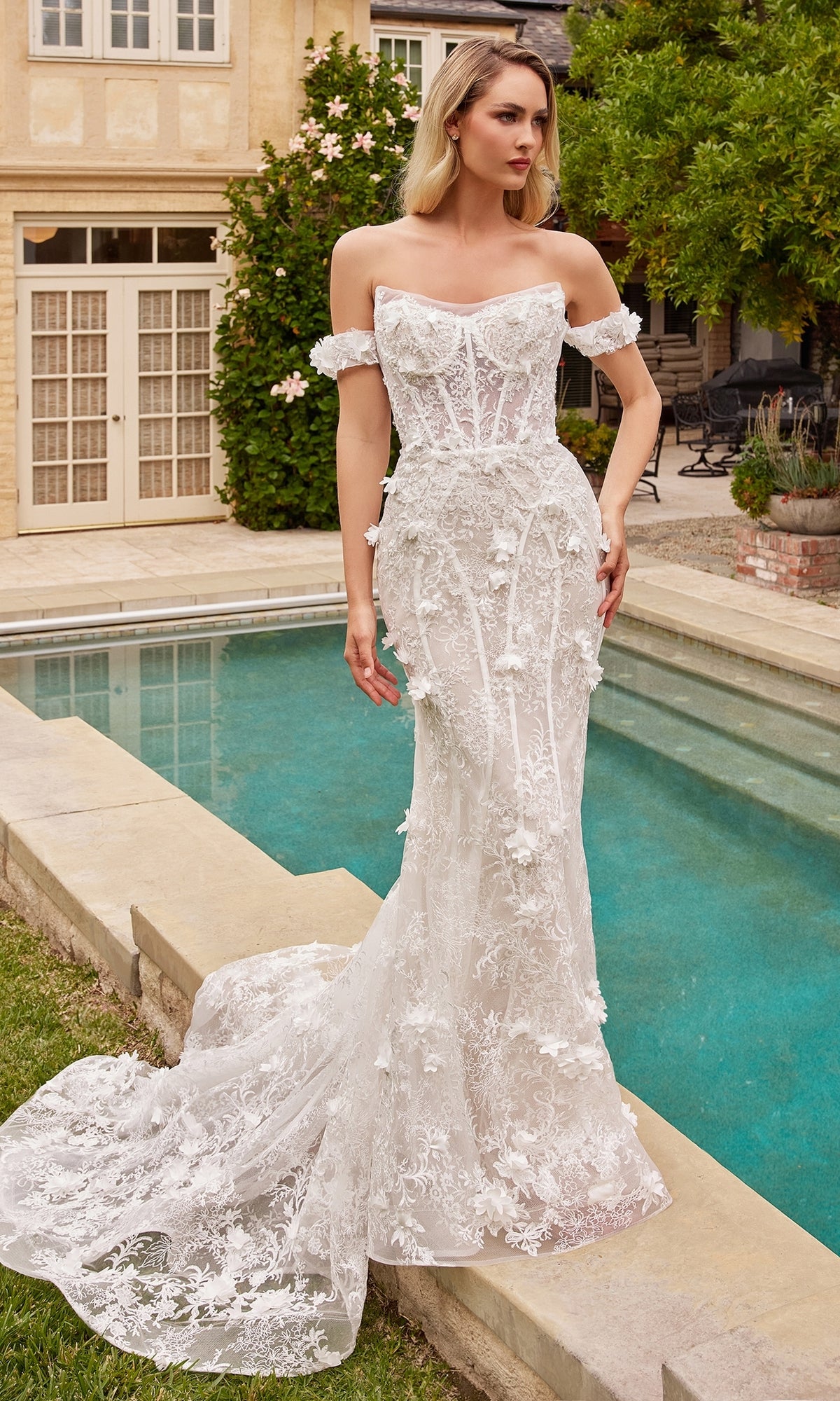A woman in the 3D Floral Lace Wedding Gown CD861W, featuring off-white lace, floral appliqués, and a removable tulle overskirt, stands by a pool in a garden.