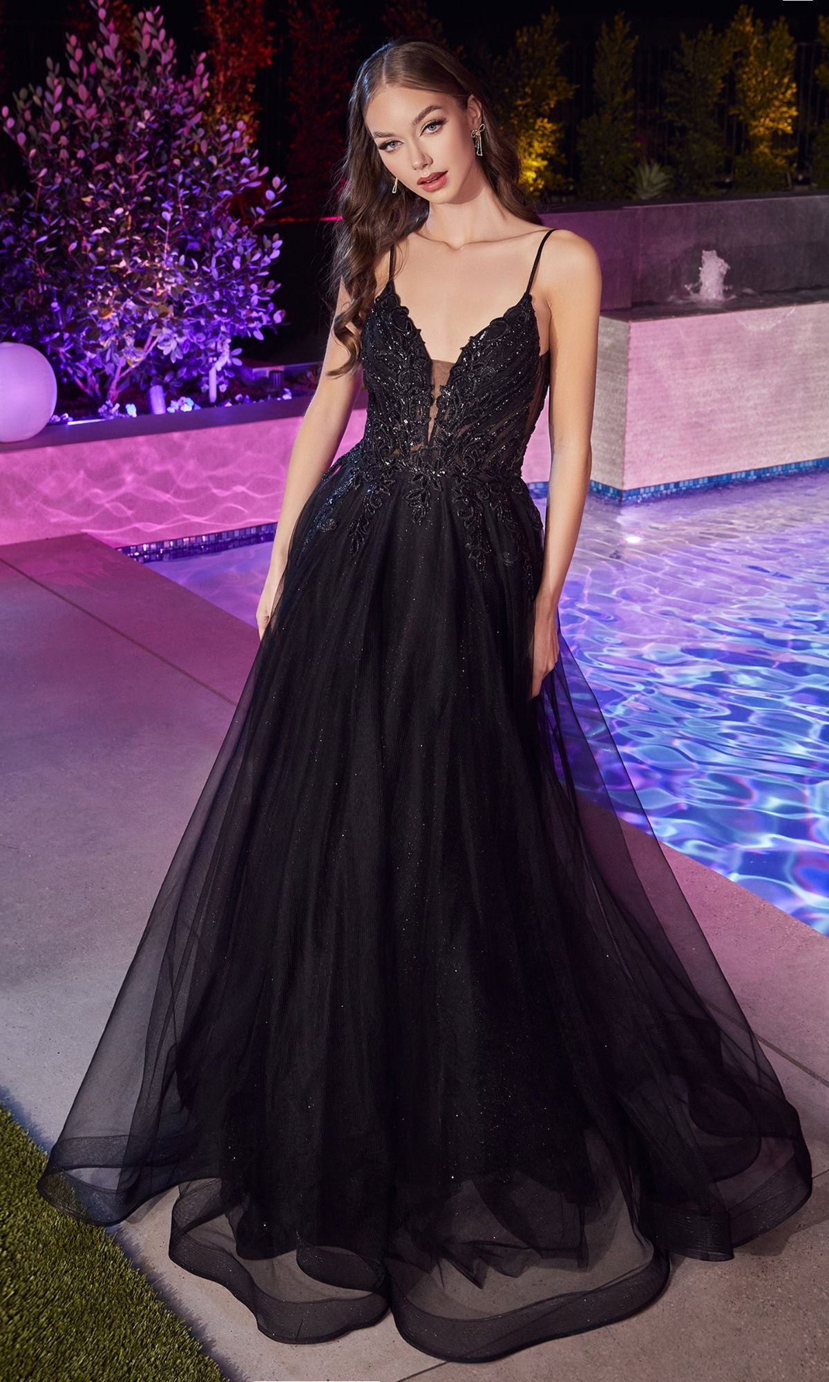 A woman wearing the Glitter-Bodice Long Prom Ball Gown CD874—a black, floor-length dress with glitter accents and a deep V-neckline—stands by a pool at night, amidst colorful lights and lush greenery.