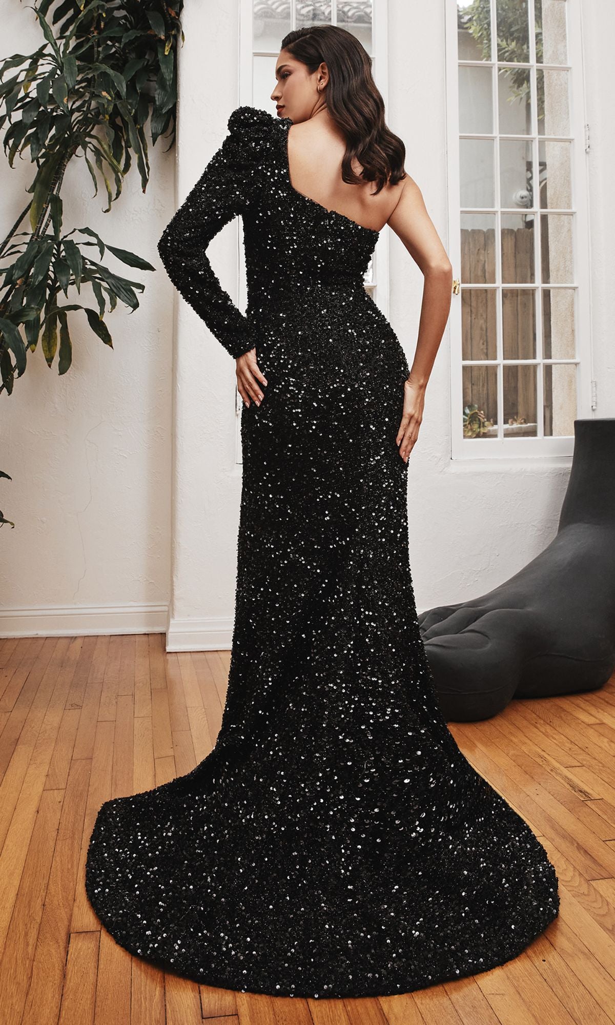 A woman stands indoors on a wooden floor, confidently posing with one hand on her hip in the One-Sleeve Long Black Sequin Formal Dress CD885, featuring dramatic sequins, a single shoulder, and an elegant train.