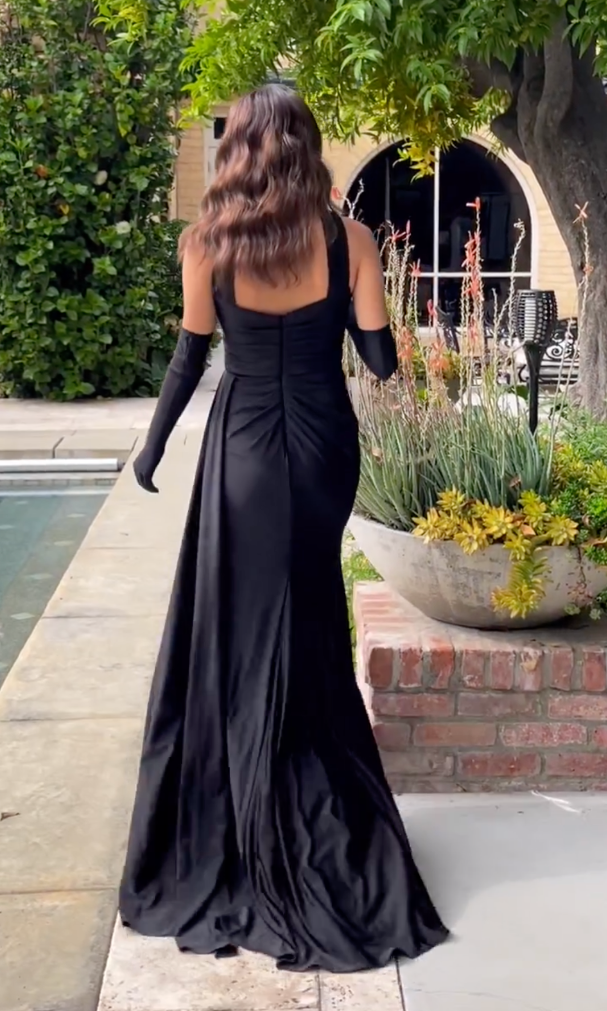A woman in the Choker Long Prom Dress with Gloves CD886 walks by a pool, surrounded by lush greenery and a large potted plant.